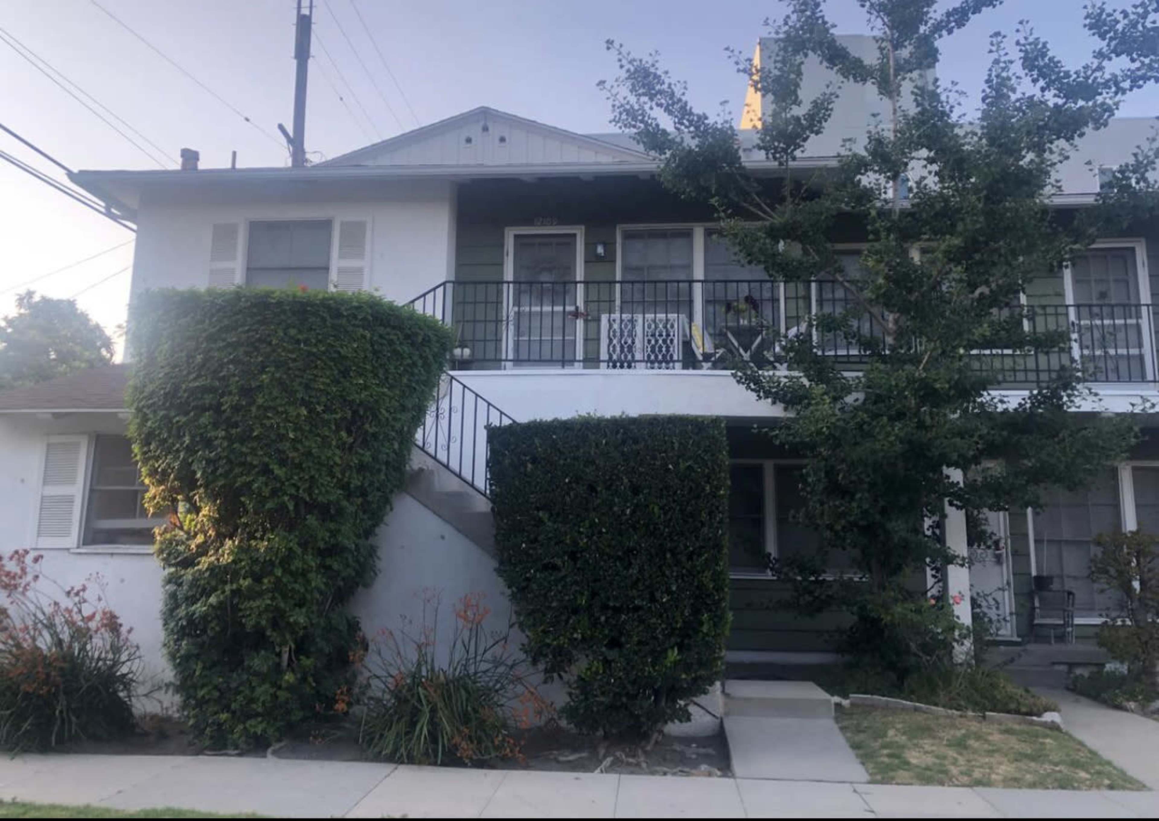 A two-story residential building features a front staircase, a balcony, and well-maintained shrubs and landscaping.