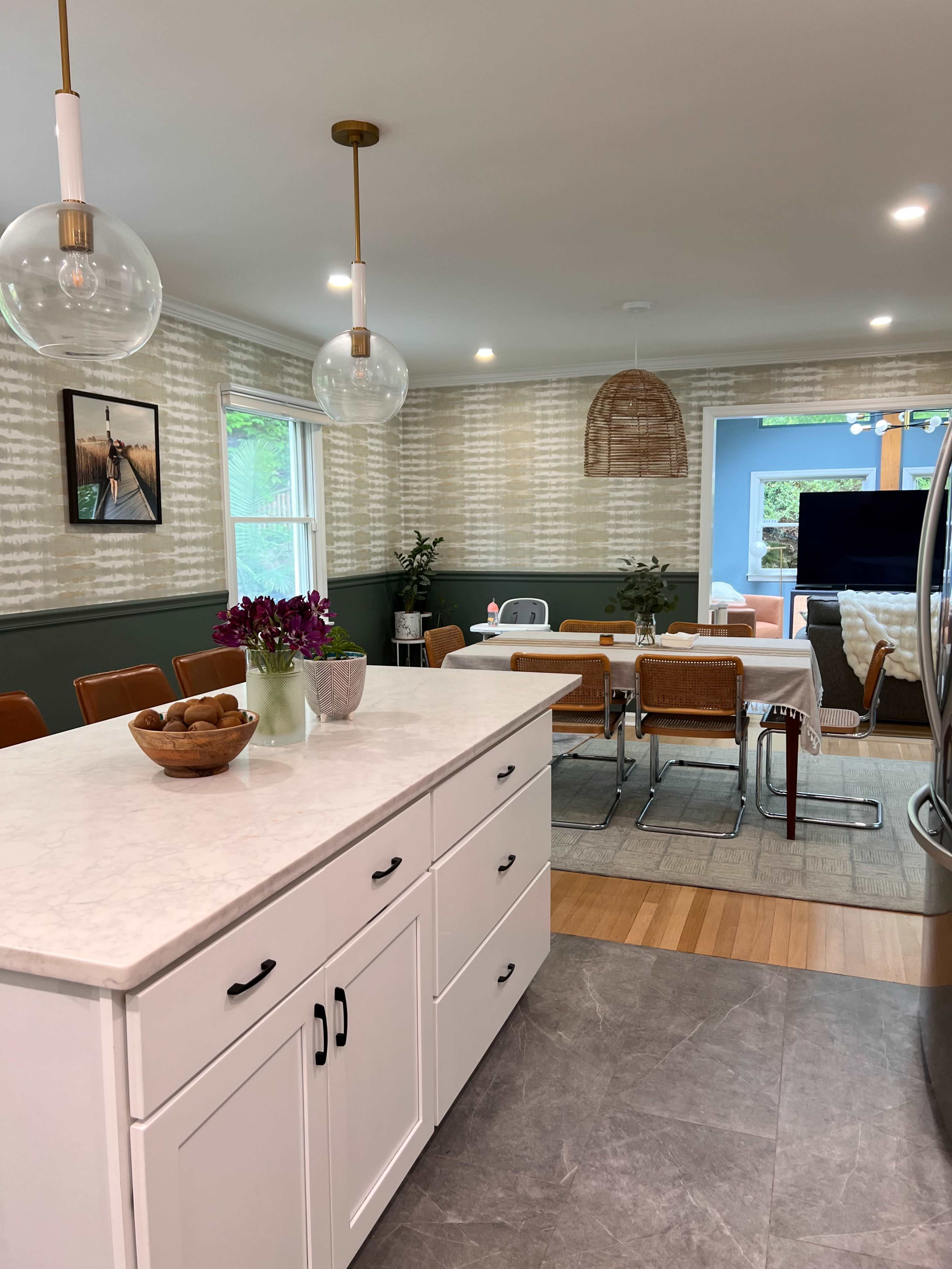 The image shows a modern kitchen with a central island, a dining area with chairs, and decorative plants against a patterned wallpaper backdrop.