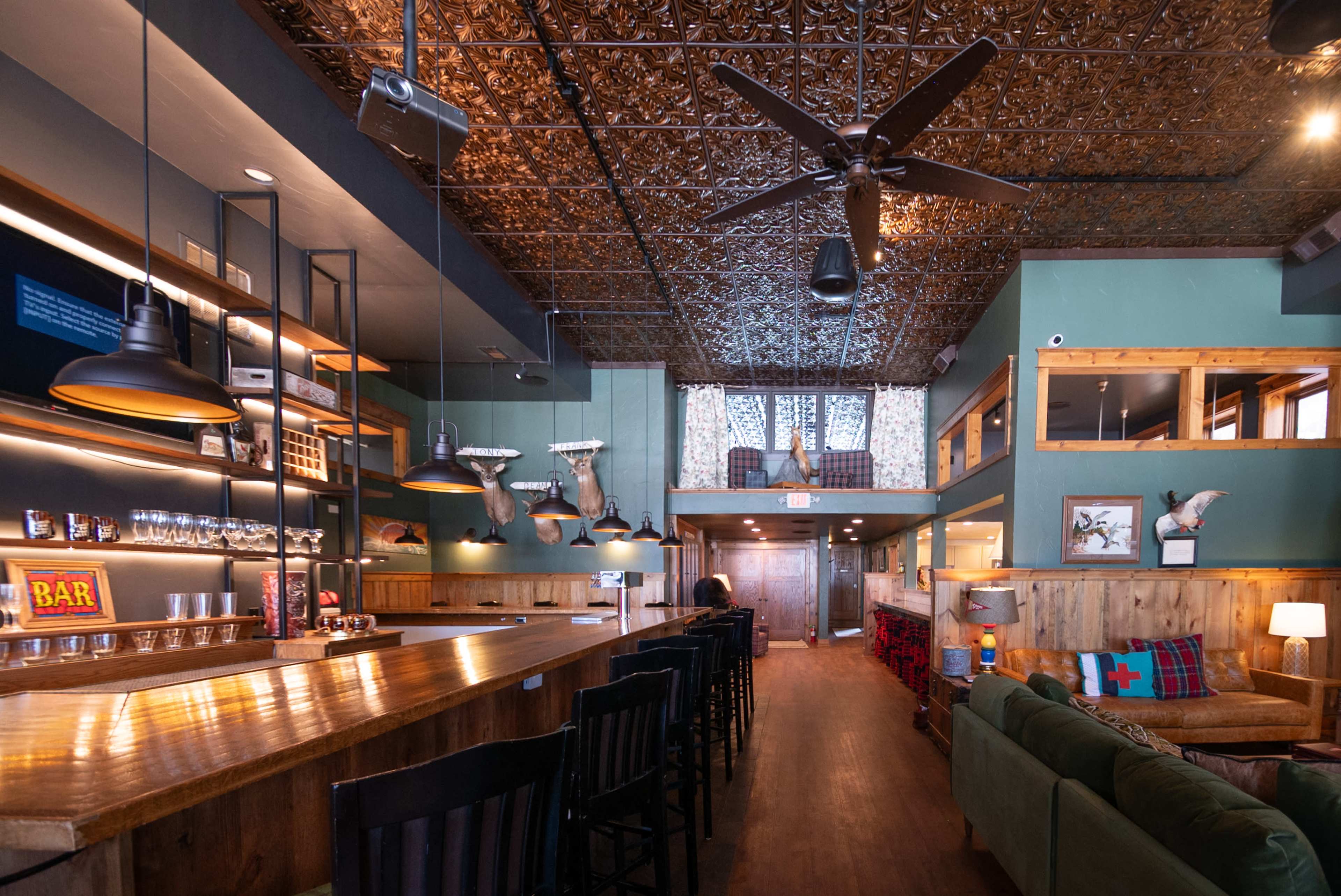 The interior of a rustic bar features wooden furnishings, a long counter, and a decorative ceiling, with comfortable seating and a cozy atmosphere.