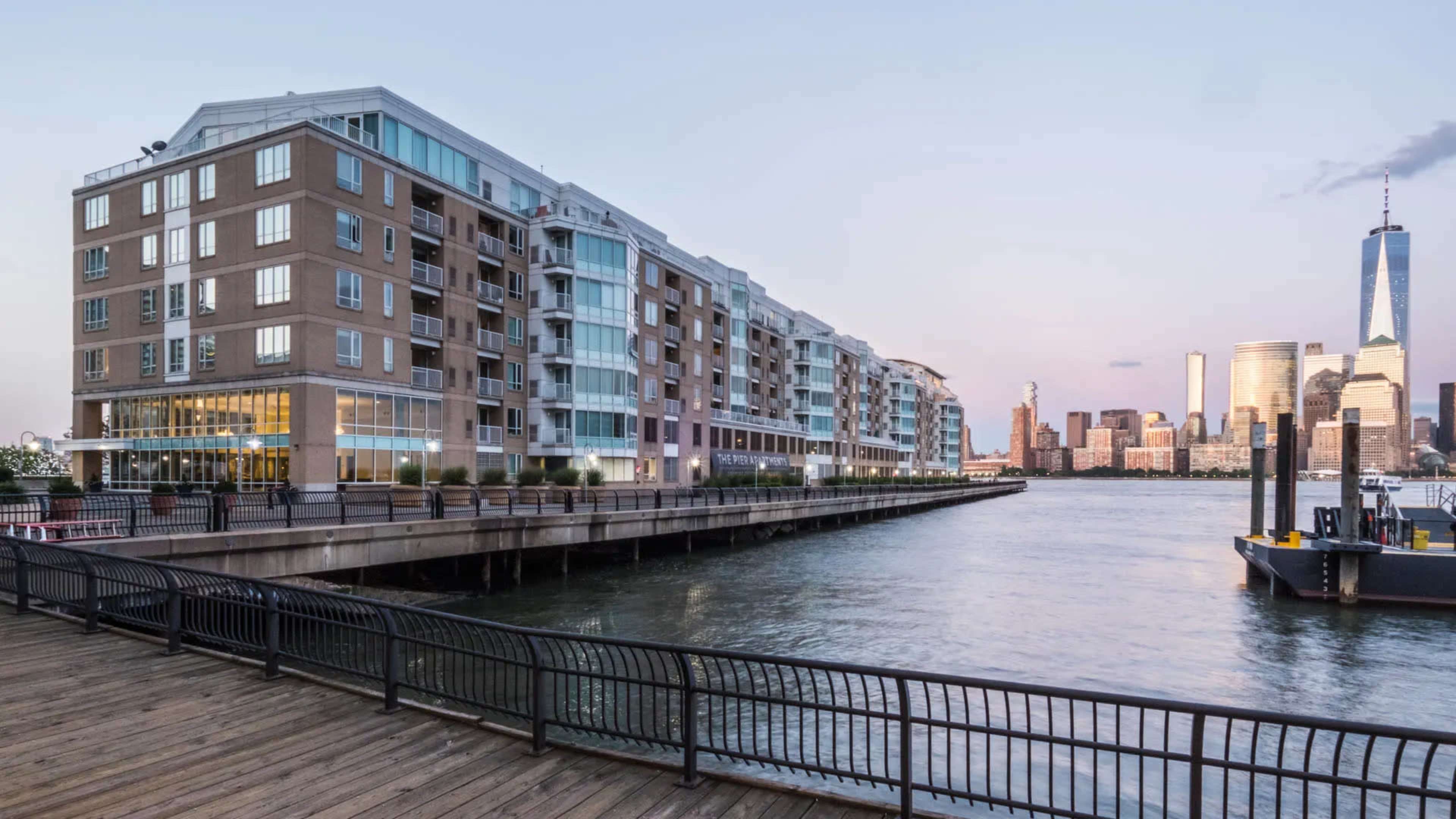 The image shows a waterfront view of a multi-story residential building adjacent to a pier with the skyline of a city in the background.