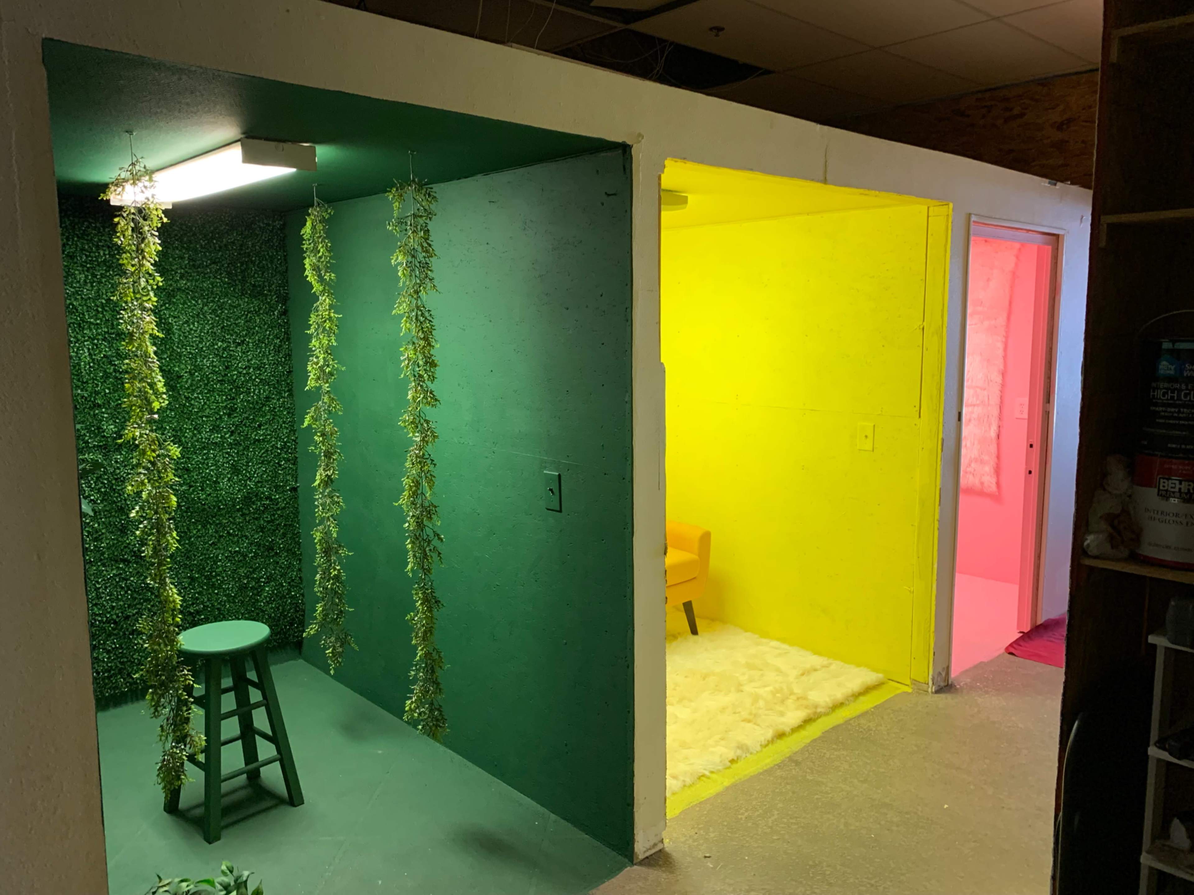 The image shows a divided room with three colored sections: a green area with a stool and hanging greenery, a bright yellow corner, and a pink space featuring a chair.