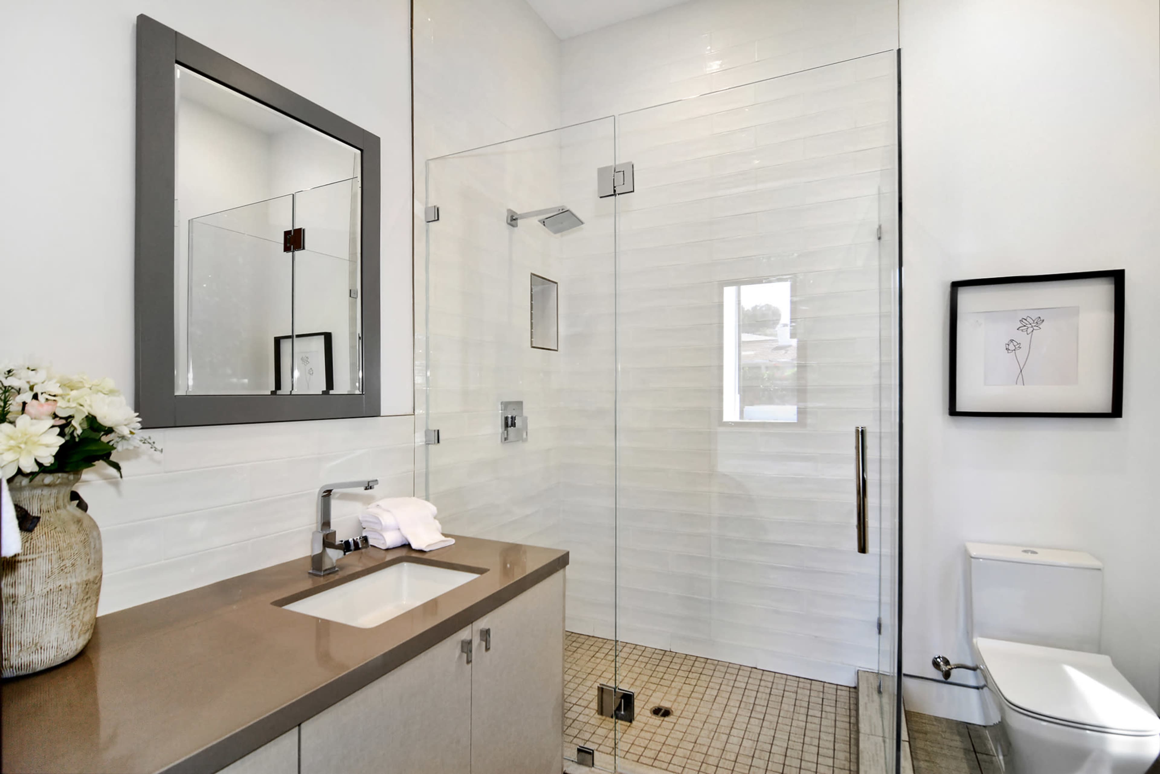 The image shows a modern bathroom with a glass shower, a rectangular mirror, a vase of flowers, and a toilet beside a sink with countertop.