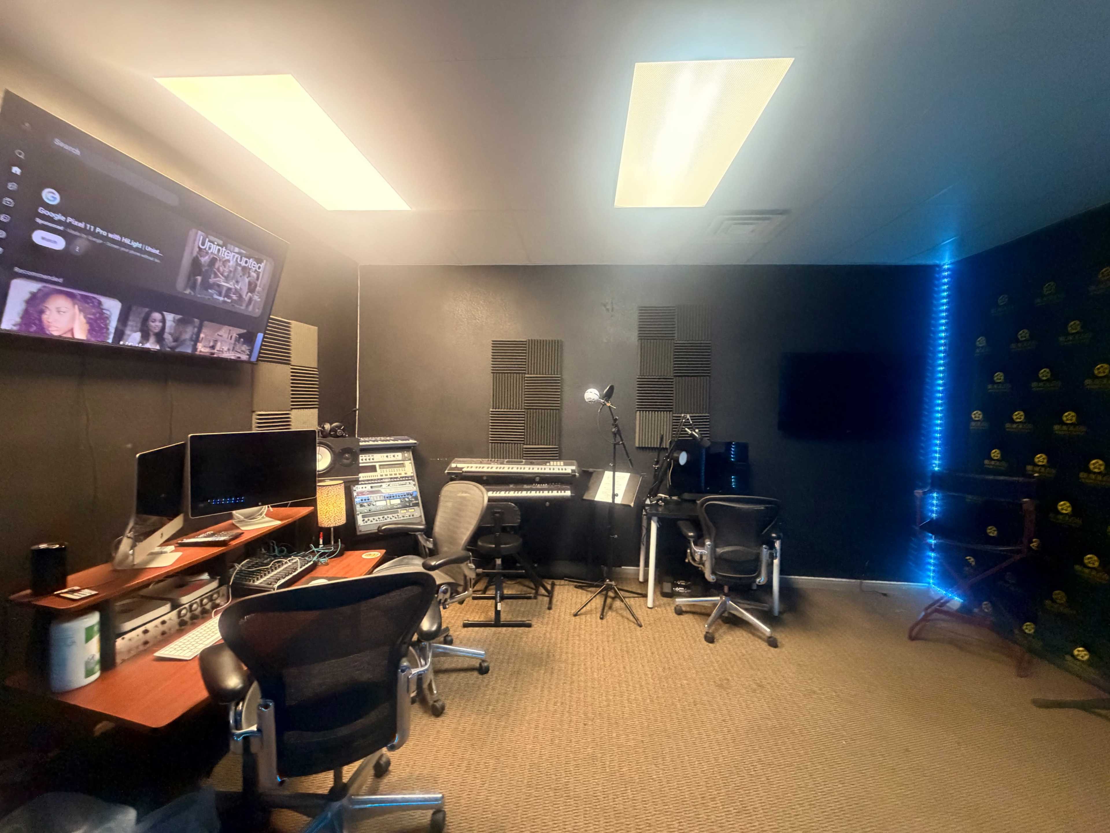 The image shows a music studio featuring a desk with recording equipment, a keyboard, and multiple chairs in a compact space.
