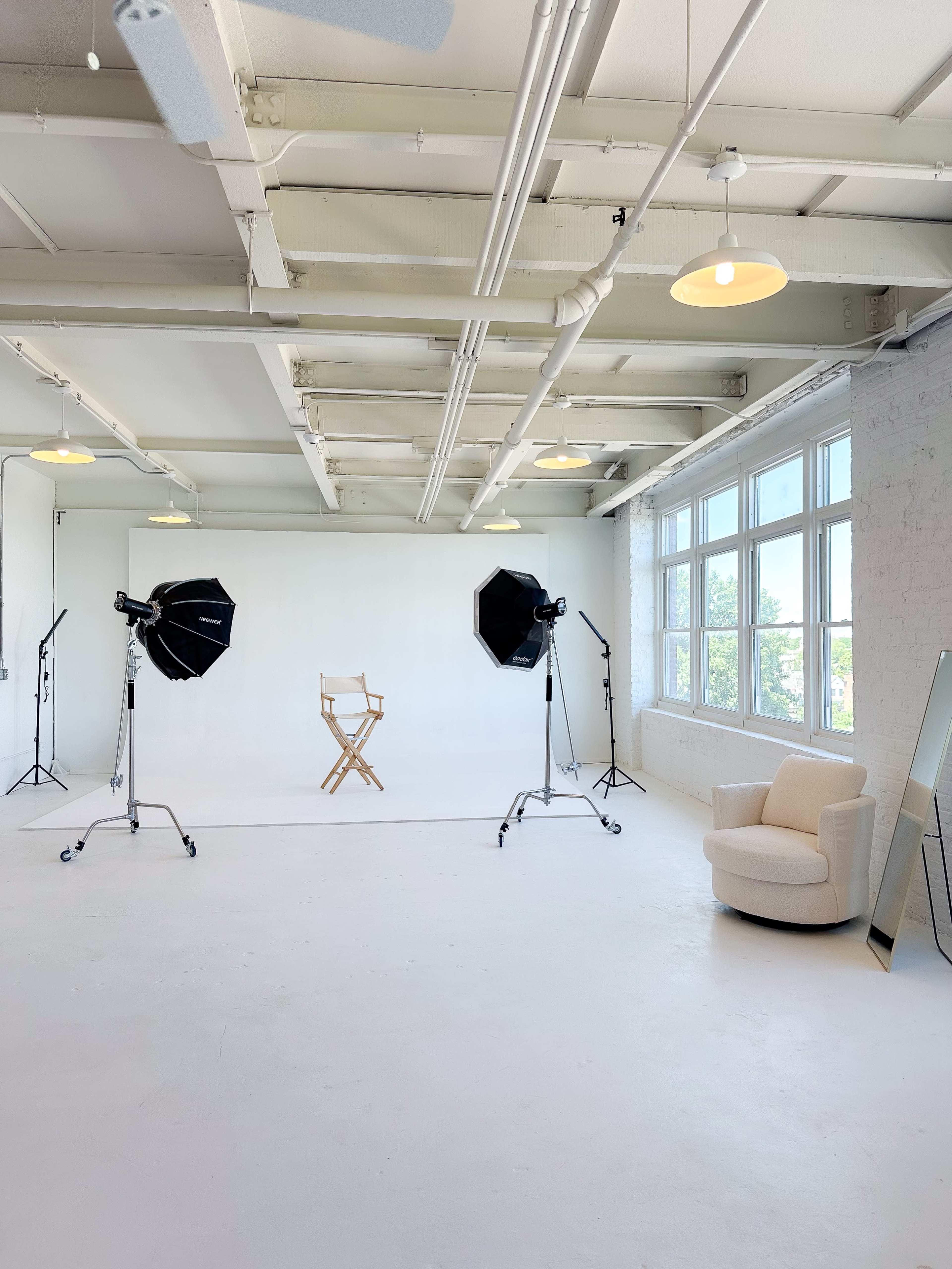 A bright, spacious photography studio features a wooden director's chair against a plain backdrop, flanked by two lighting setups and a mirror.