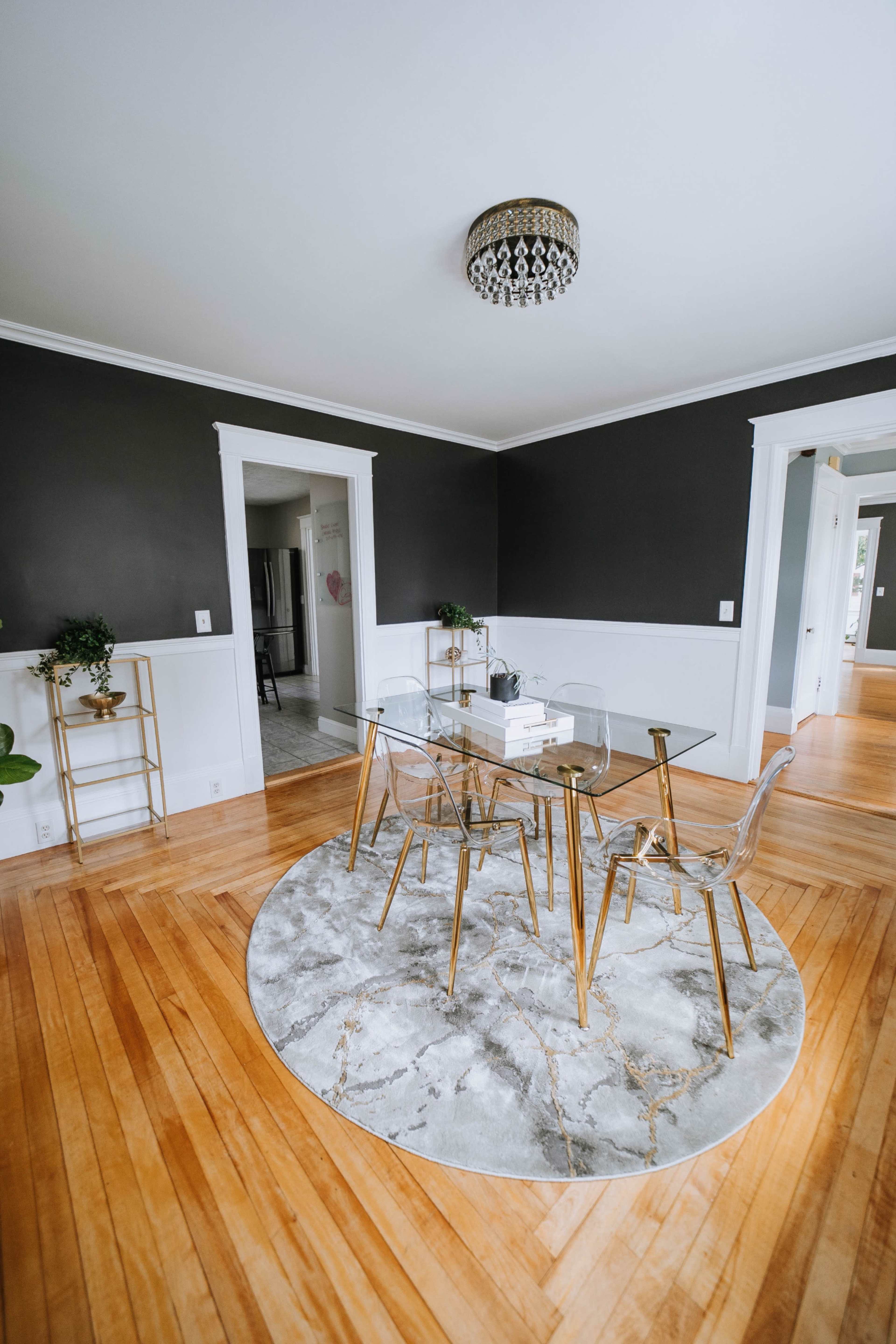 A clear glass dining table with gold legs is surrounded by transparent chairs on a marble-patterned round rug in a room with dark walls and hardwood floors.