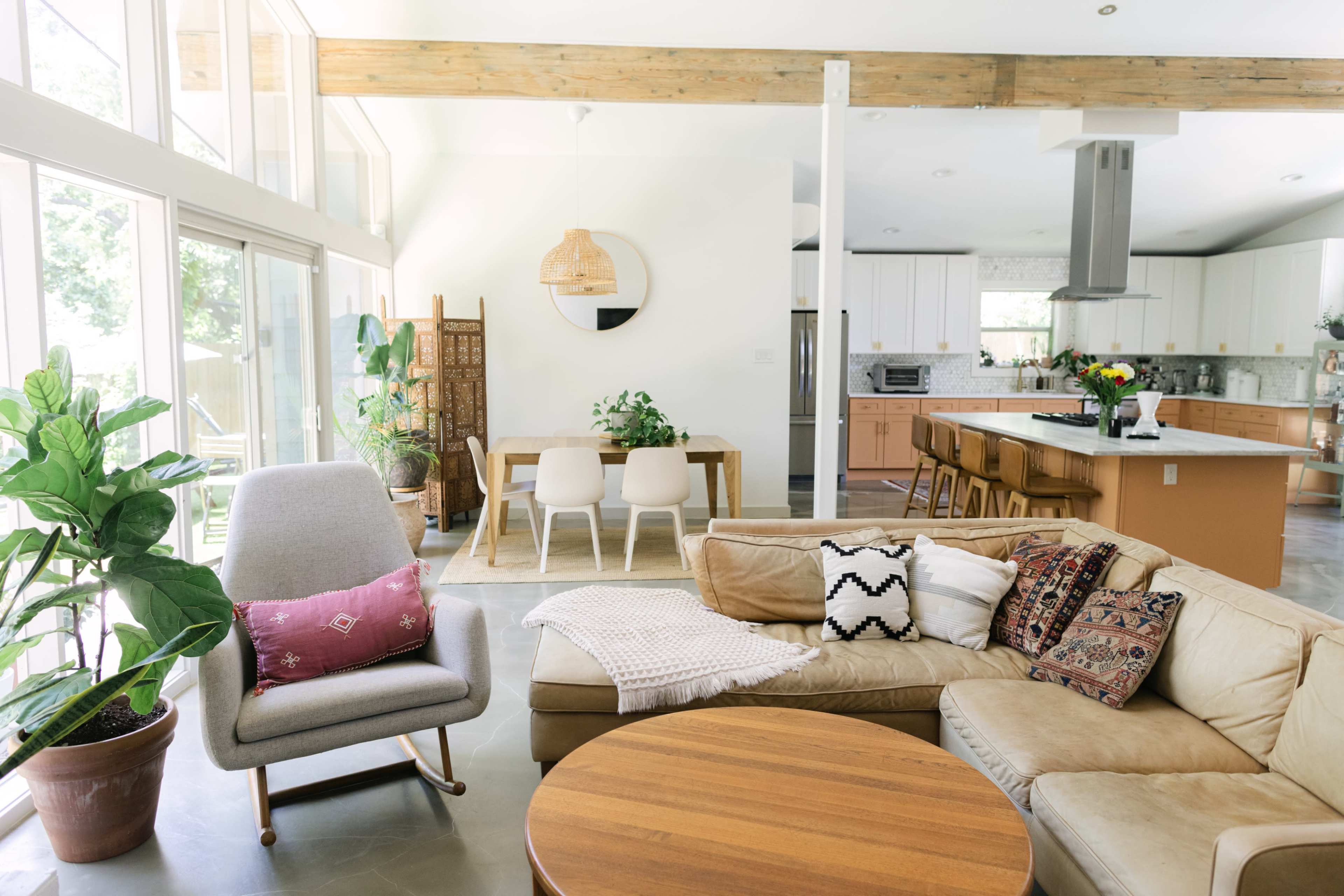 A spacious living area features a beige sectional sofa, a round wooden table, and large windows allowing natural light, with plants and modern furniture included in the design.