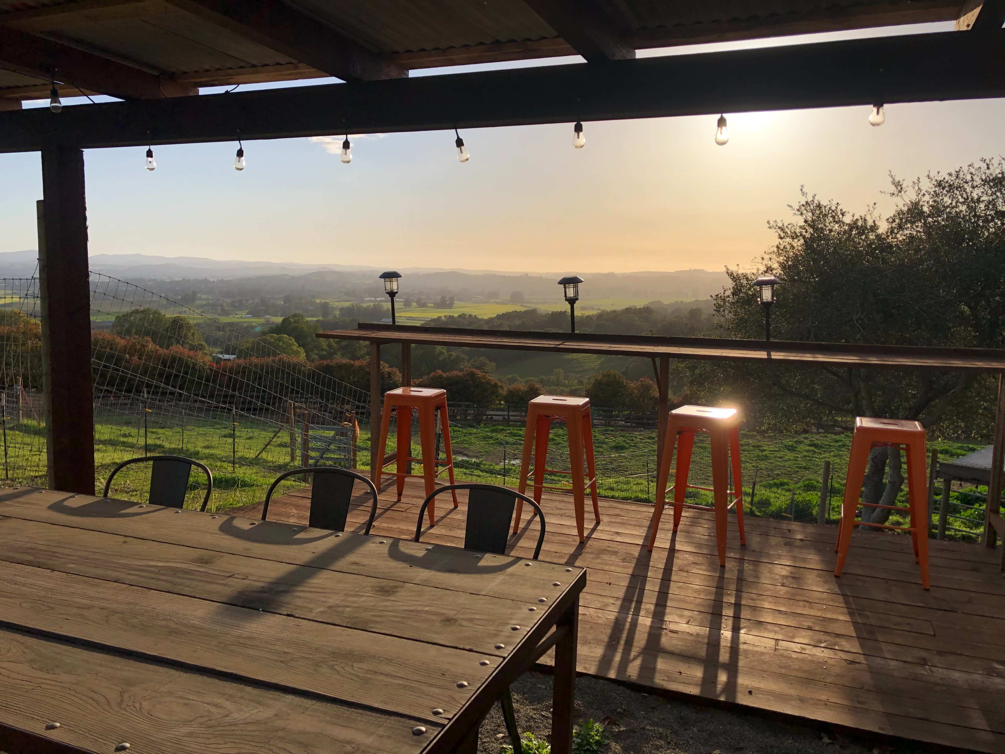 A wooden deck features tall orange stools with a scenic view of rolling hills at sunset.