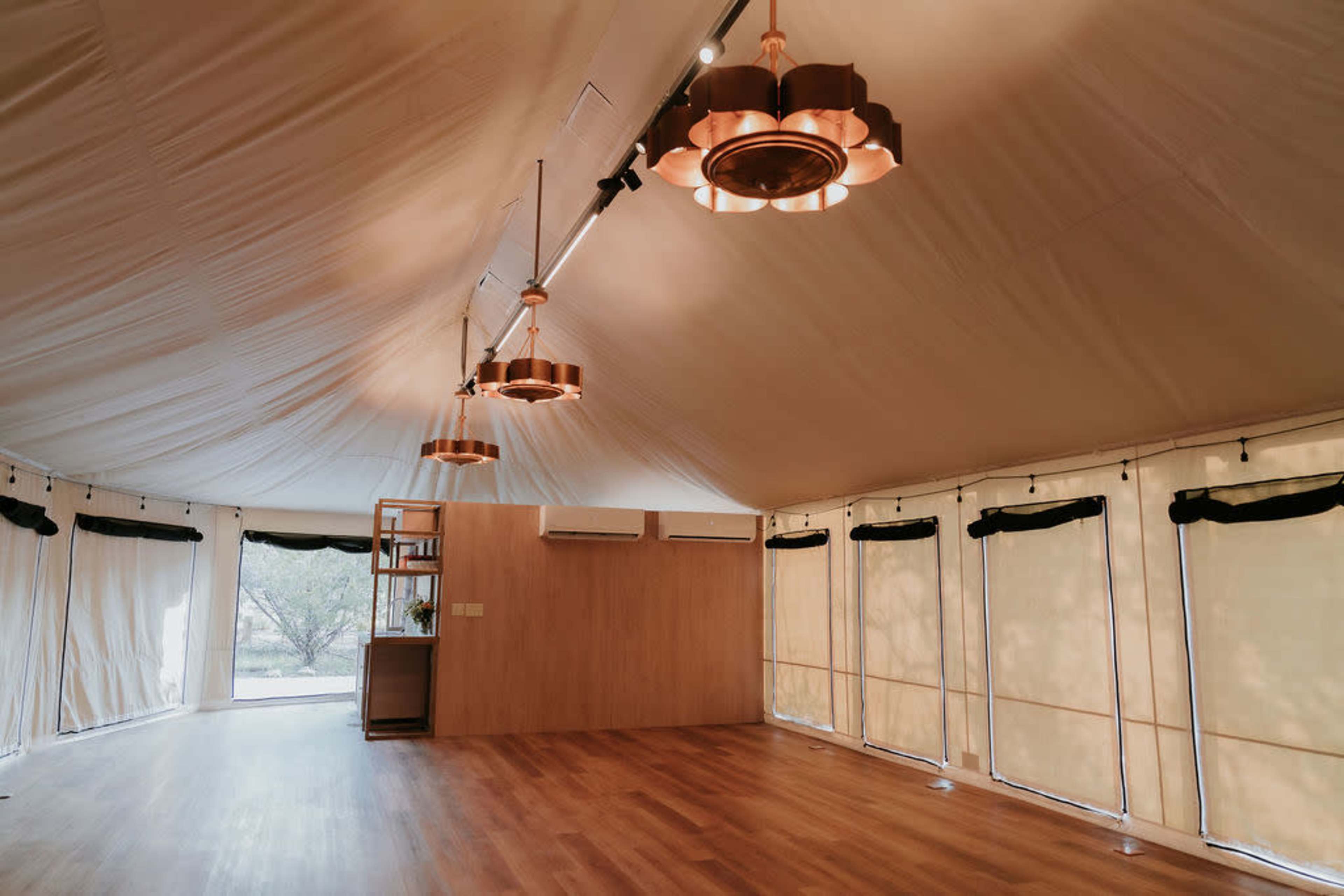 The interior of a spacious tent featuring wooden flooring, overhead lighting fixtures, and large windows with fabric coverings.