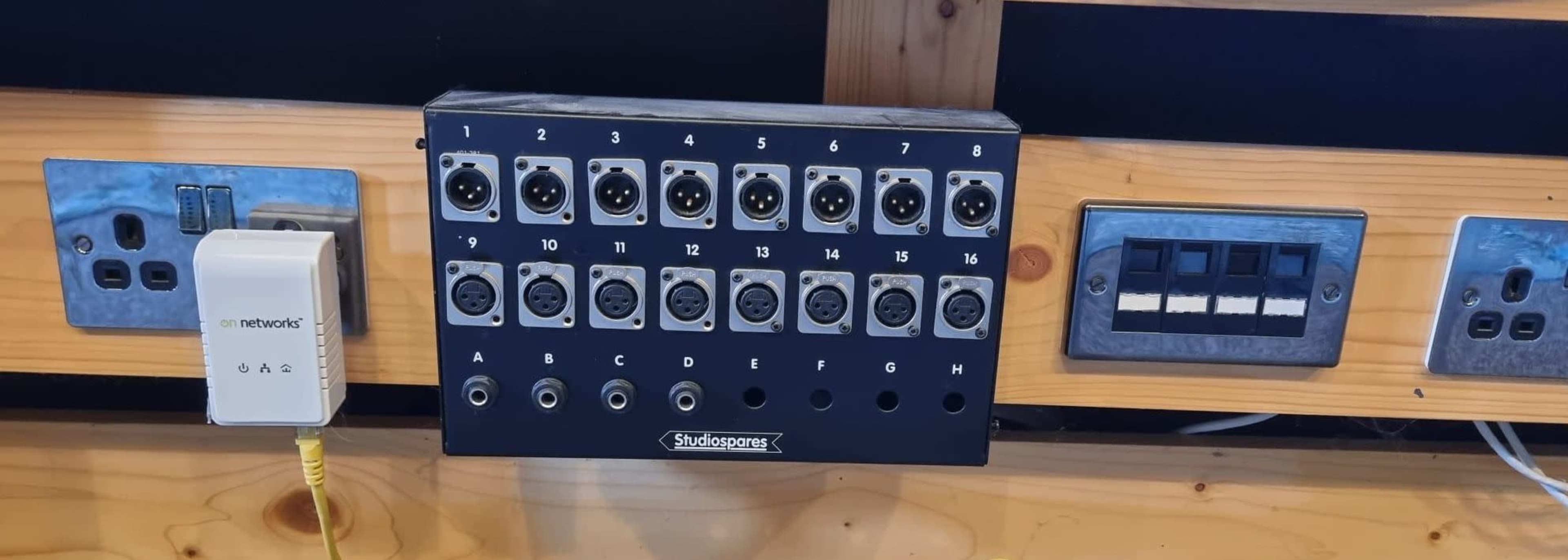 The image shows a wall-mounted audio interface with multiple XLR connectors and additional input/output ports, alongside electrical sockets and a network connection point.