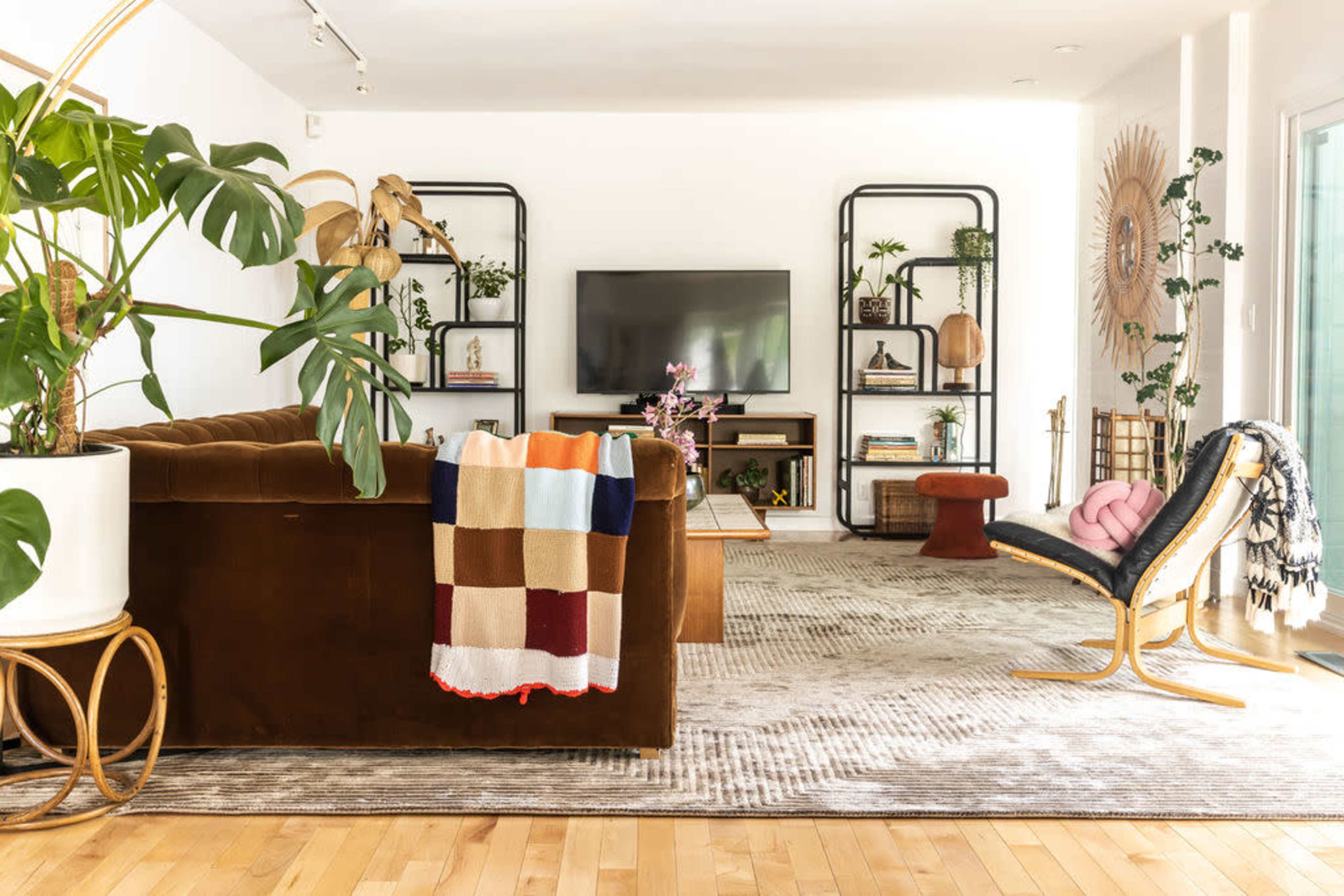 The image shows a modern living room with a brown upholstered sofa, a large television mounted on the wall, and various decorative shelves filled with plants and accessories.