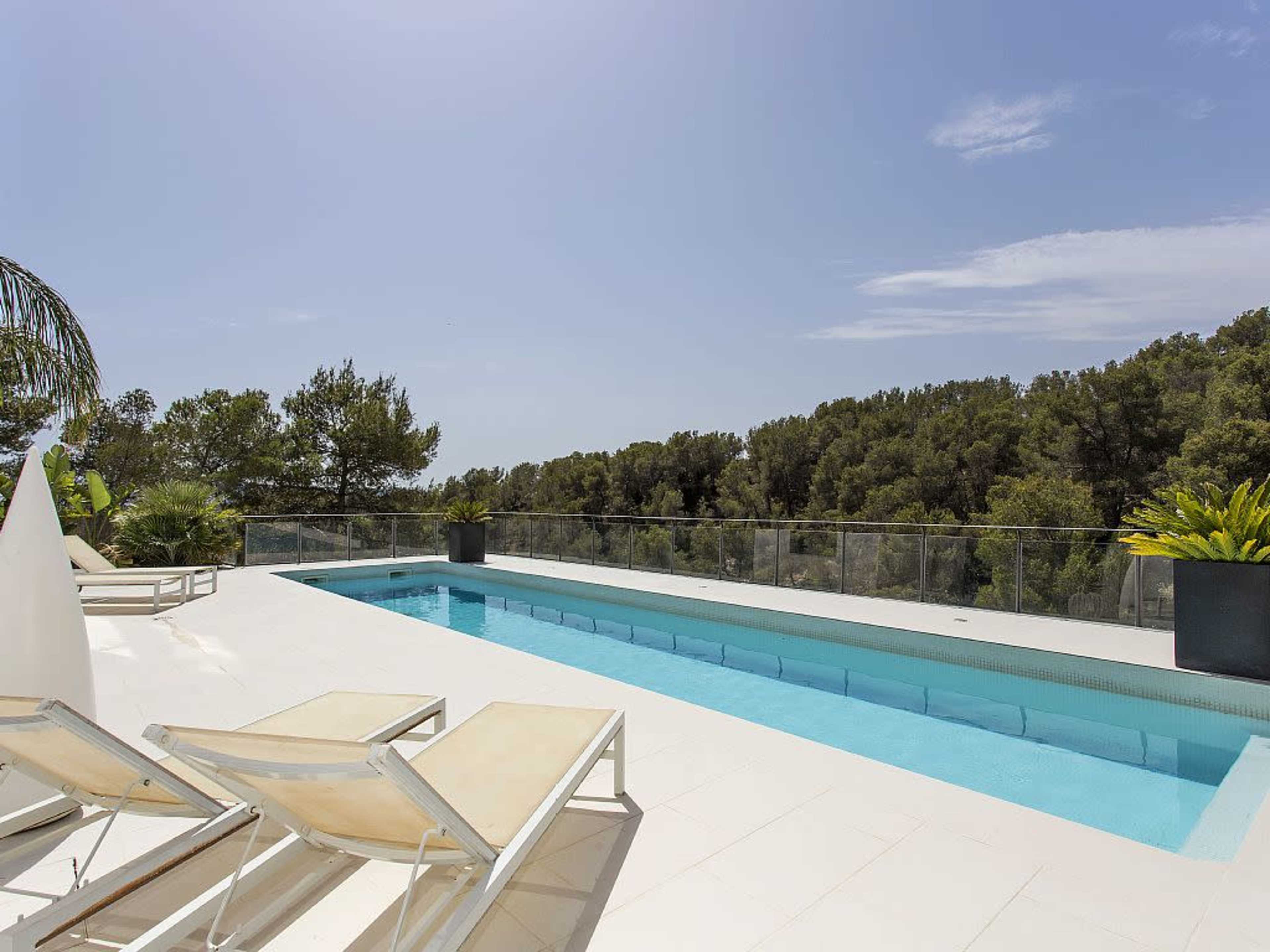 A clean, modern pool area features lounge chairs beside a rectangular swimming pool, surrounded by greenery and clear blue skies.
