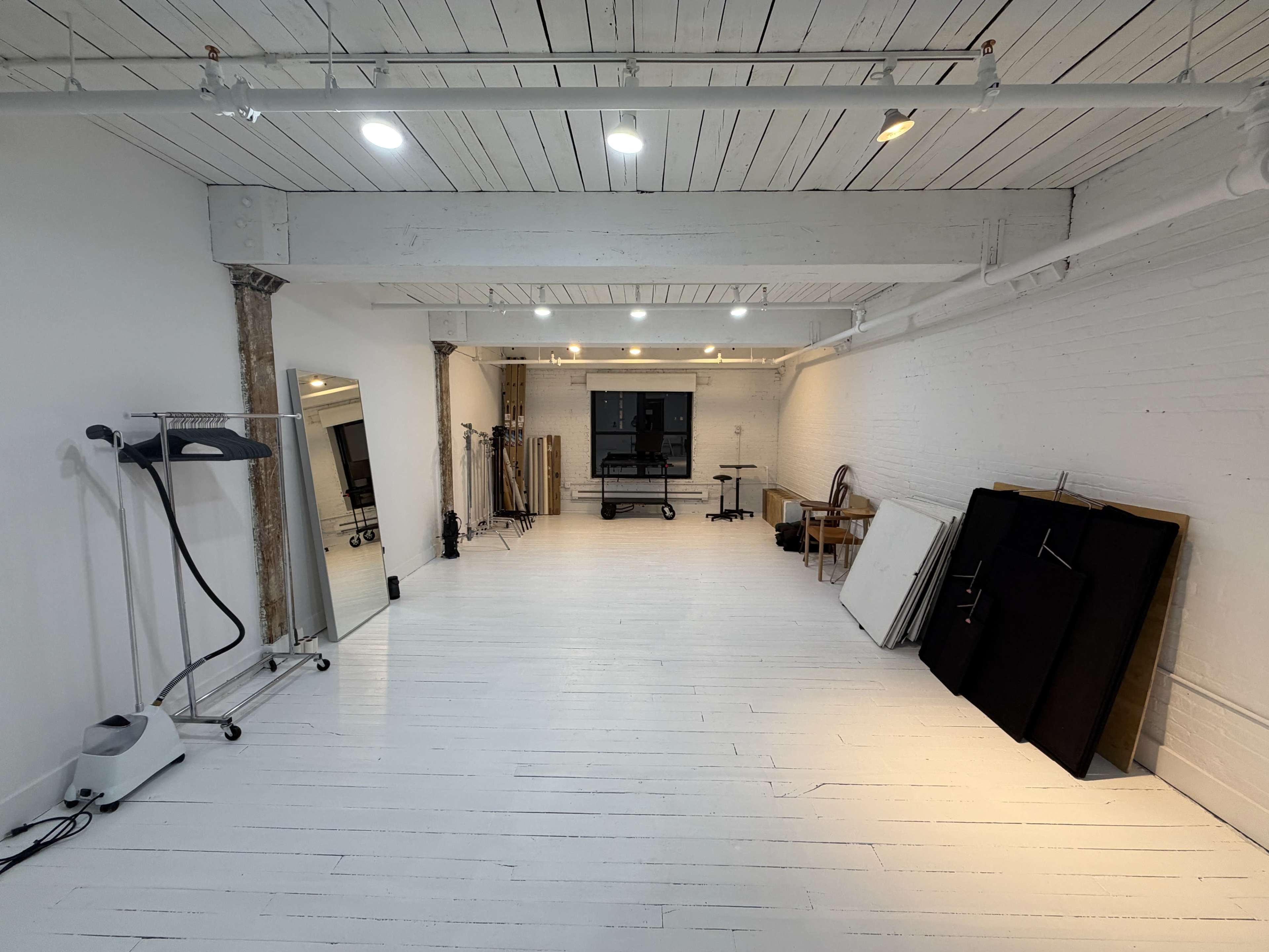 The image shows a spacious studio with white wooden floors and walls, featuring a mirror, a clothing rack, and various equipment along the sides.