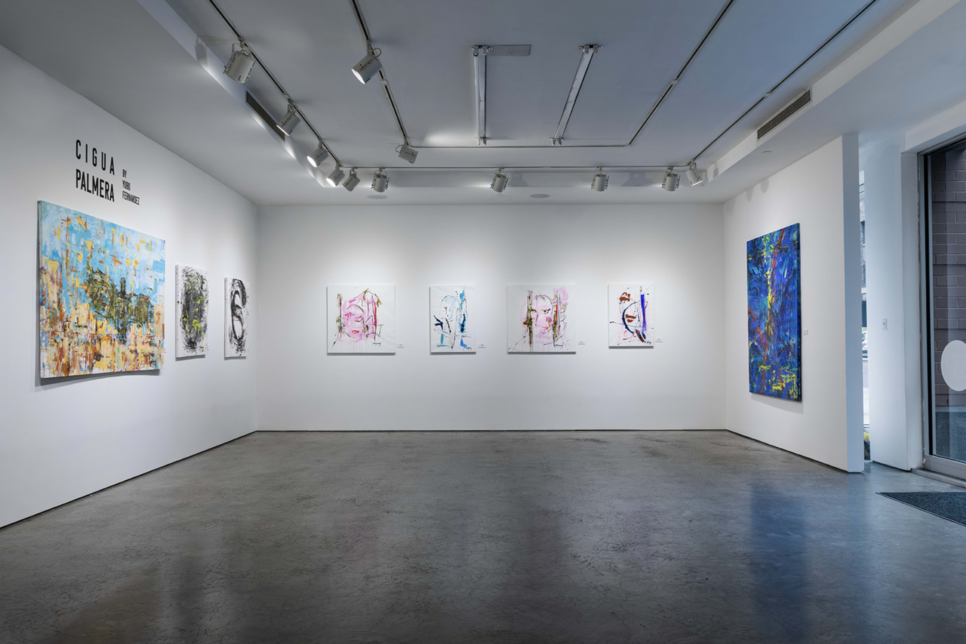 An art gallery displays a series of colorful abstract paintings on white walls, with a polished concrete floor and natural light coming from a large window.
