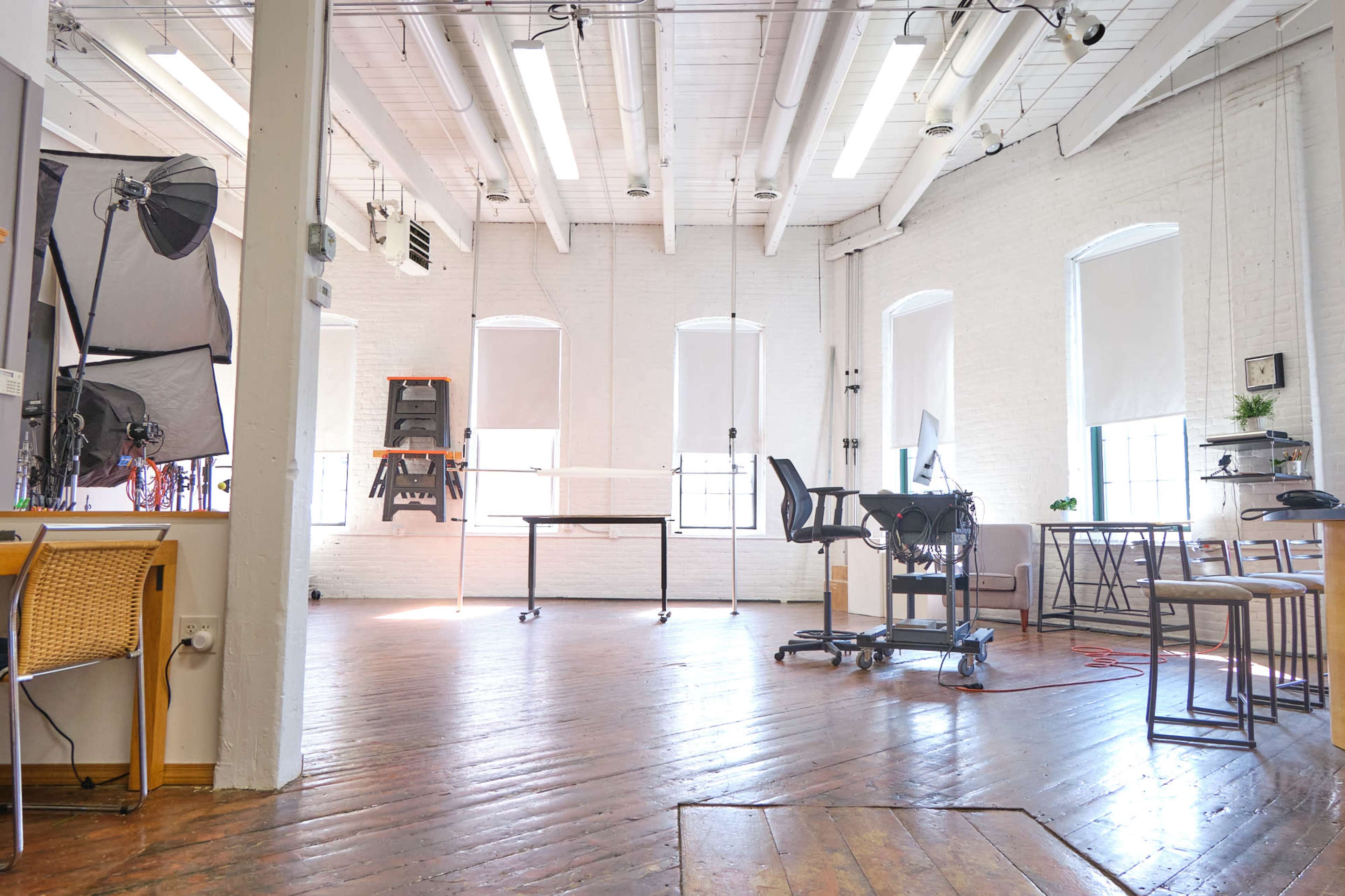 A bright, open workspace featuring wooden floors, large windows with shades, and various furniture including a desk and chairs.