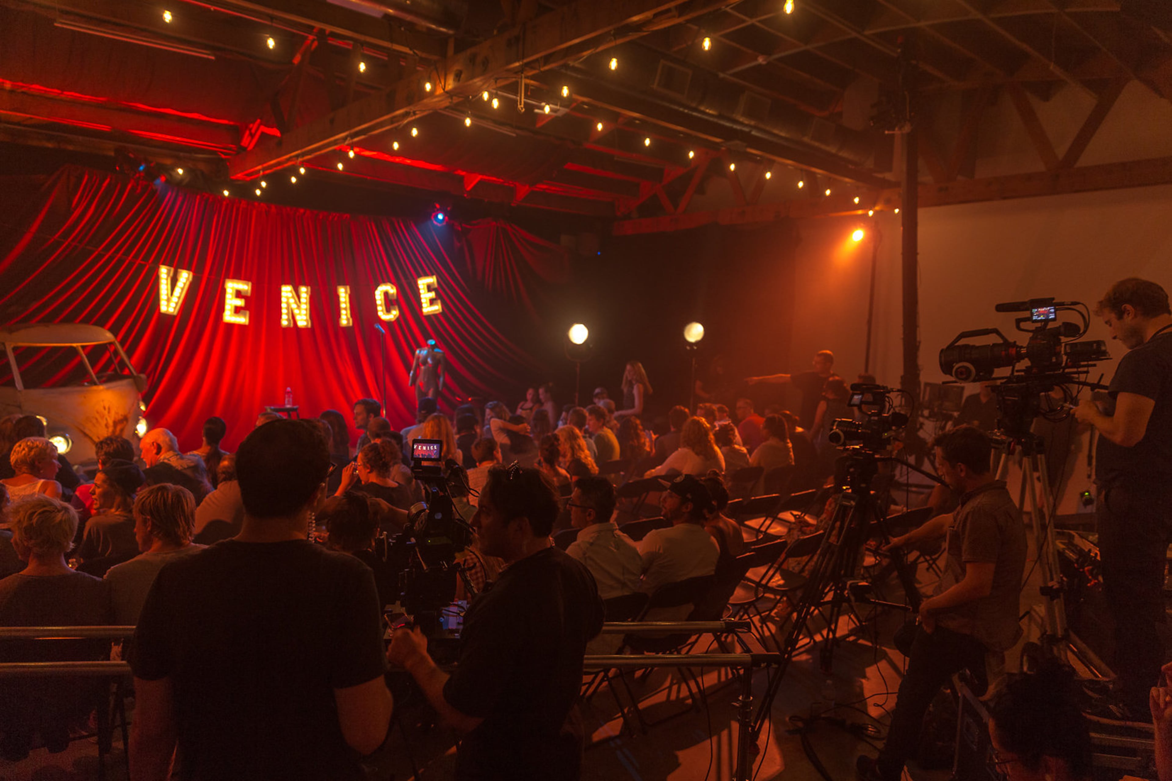 A crowd gathers in a dimly lit venue with a large "VENICE" sign illuminated on stage, while cameras capture the performance.