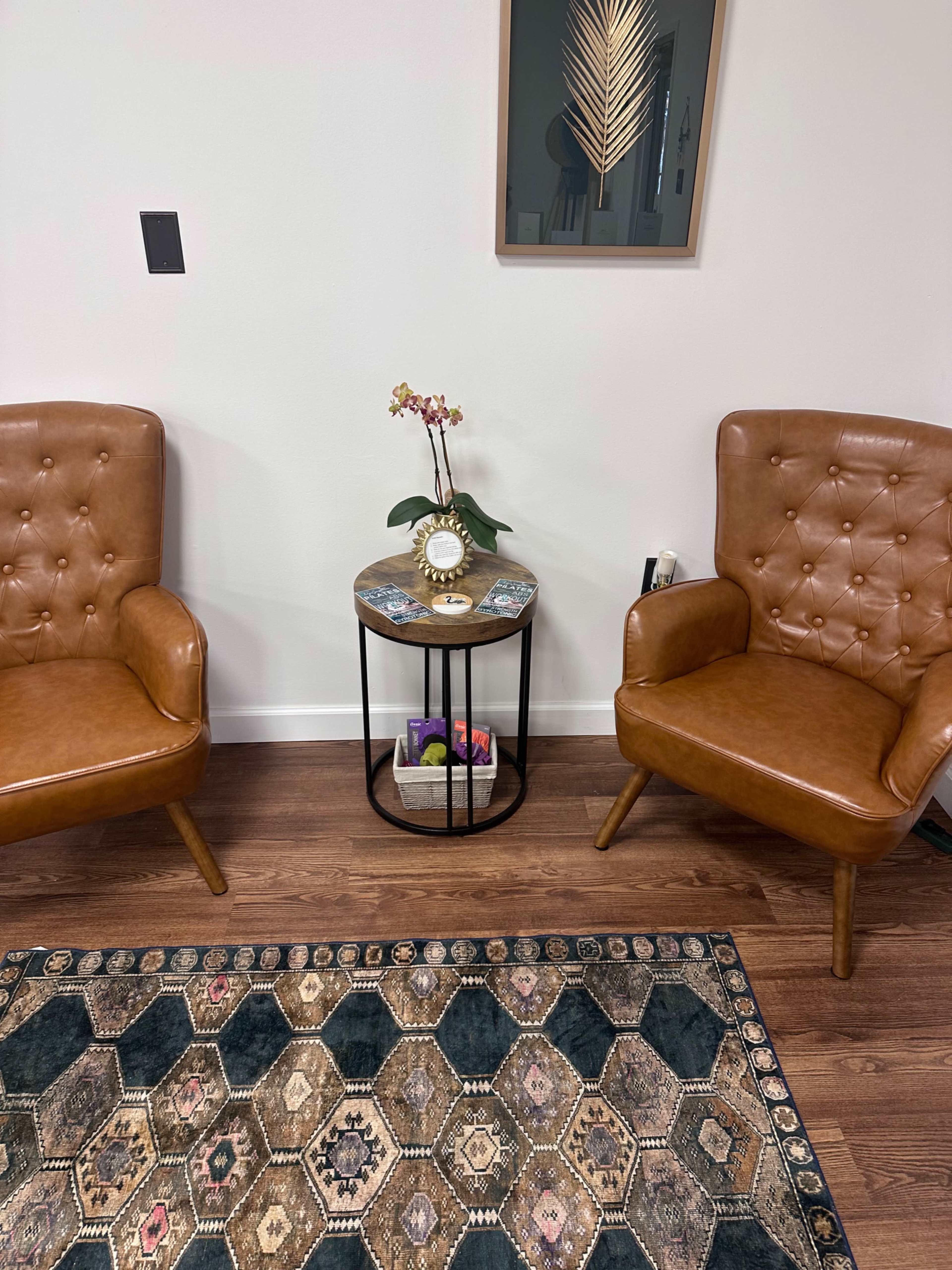 The image shows two brown tufted armchairs flanking a circular side table, which hosts a small plant and a decorative clock, all set on a patterned area rug.