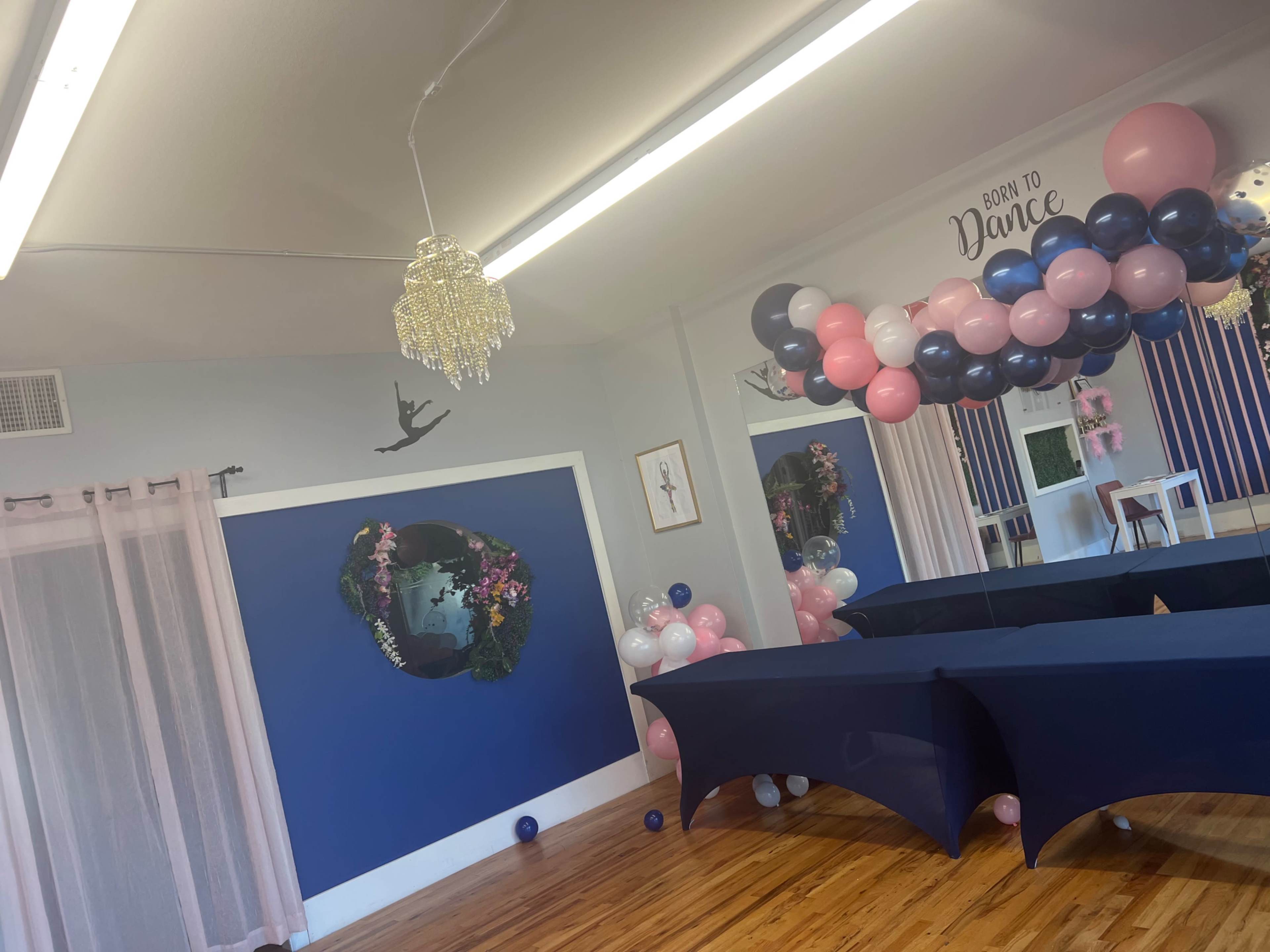 A decorated dance studio featuring blue walls, a chandelier, balloon garlands in pink and navy colors, and dark tables arranged in the space.