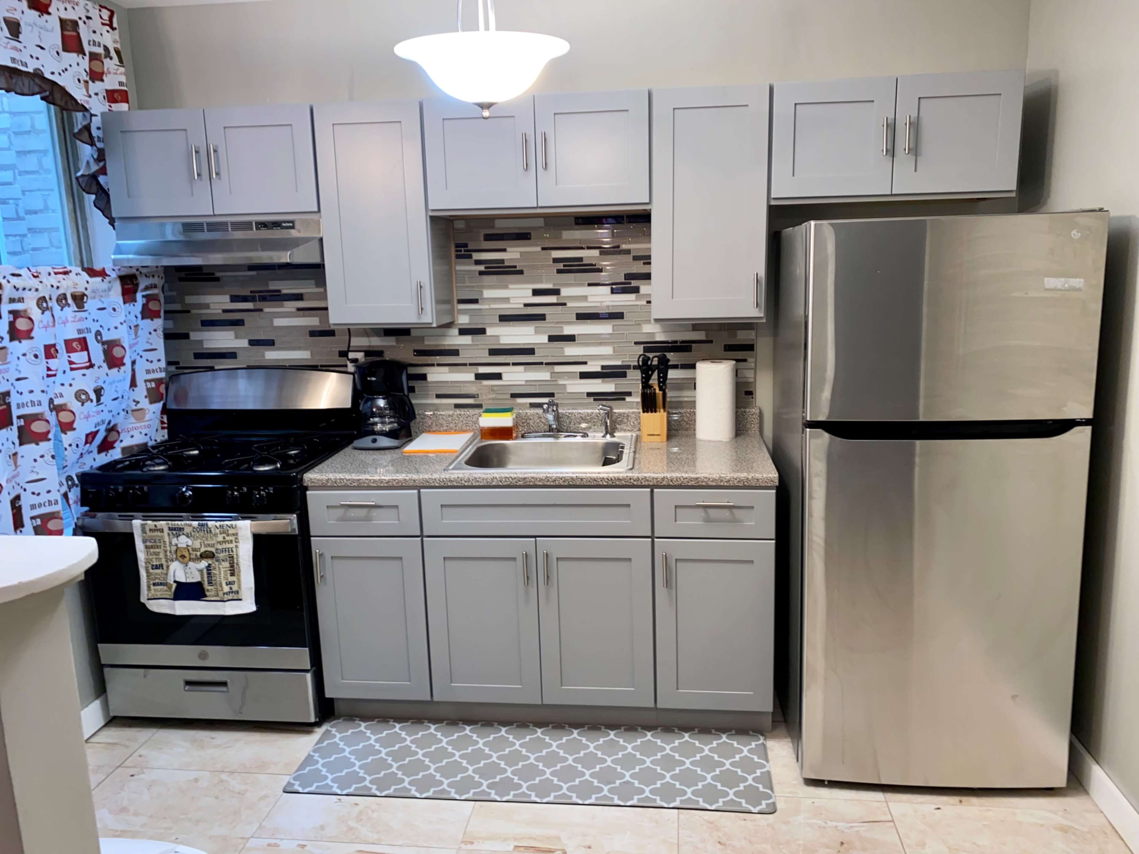 The image shows a modern kitchen featuring gray cabinets, a stainless steel refrigerator, a black gas stove, and a decorative backsplash with tiles.