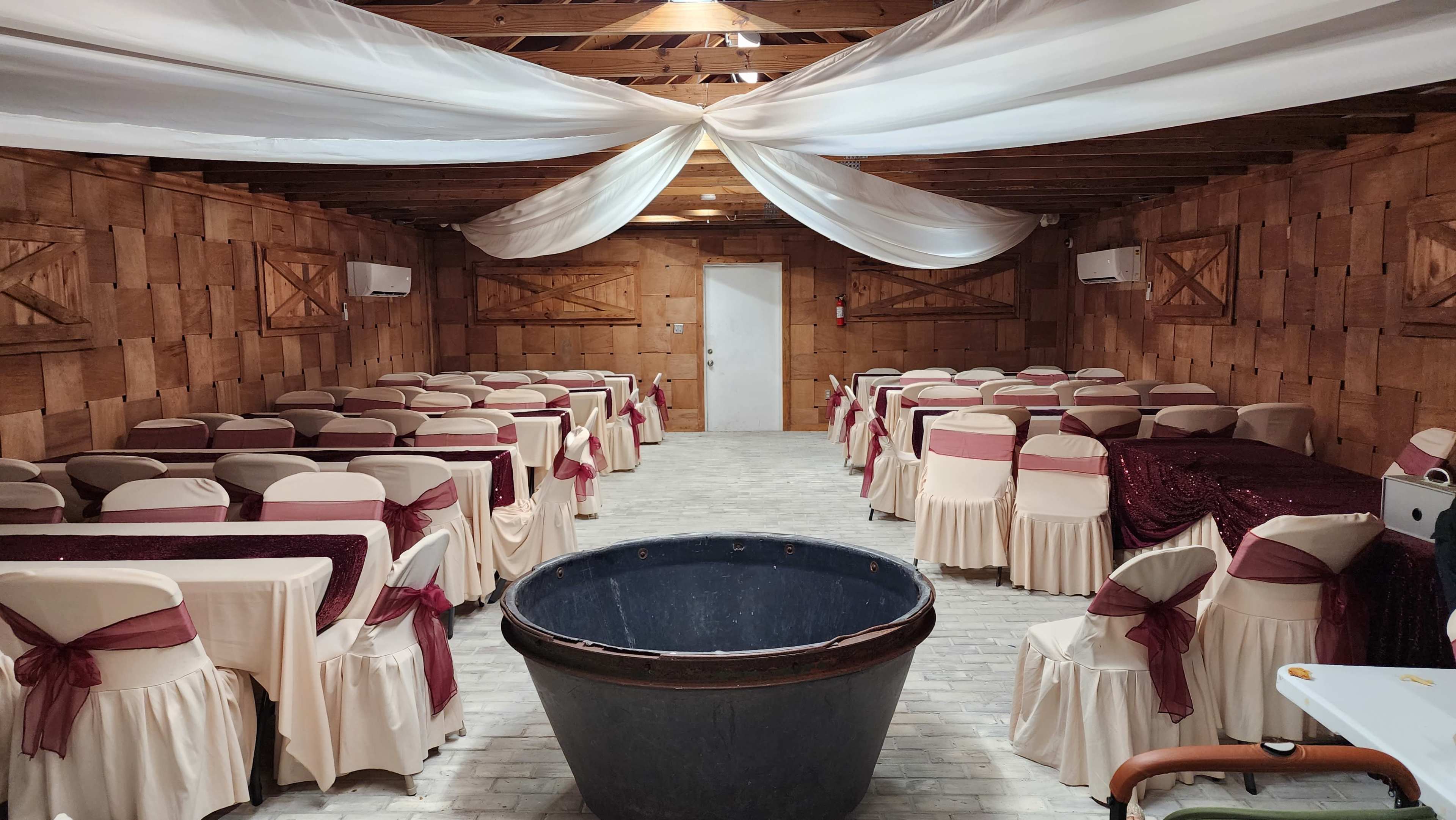 The image shows an indoor venue with rows of tables covered in white tablecloths and burgundy accents, a large black basin in the center, and draped fabric on the ceiling.