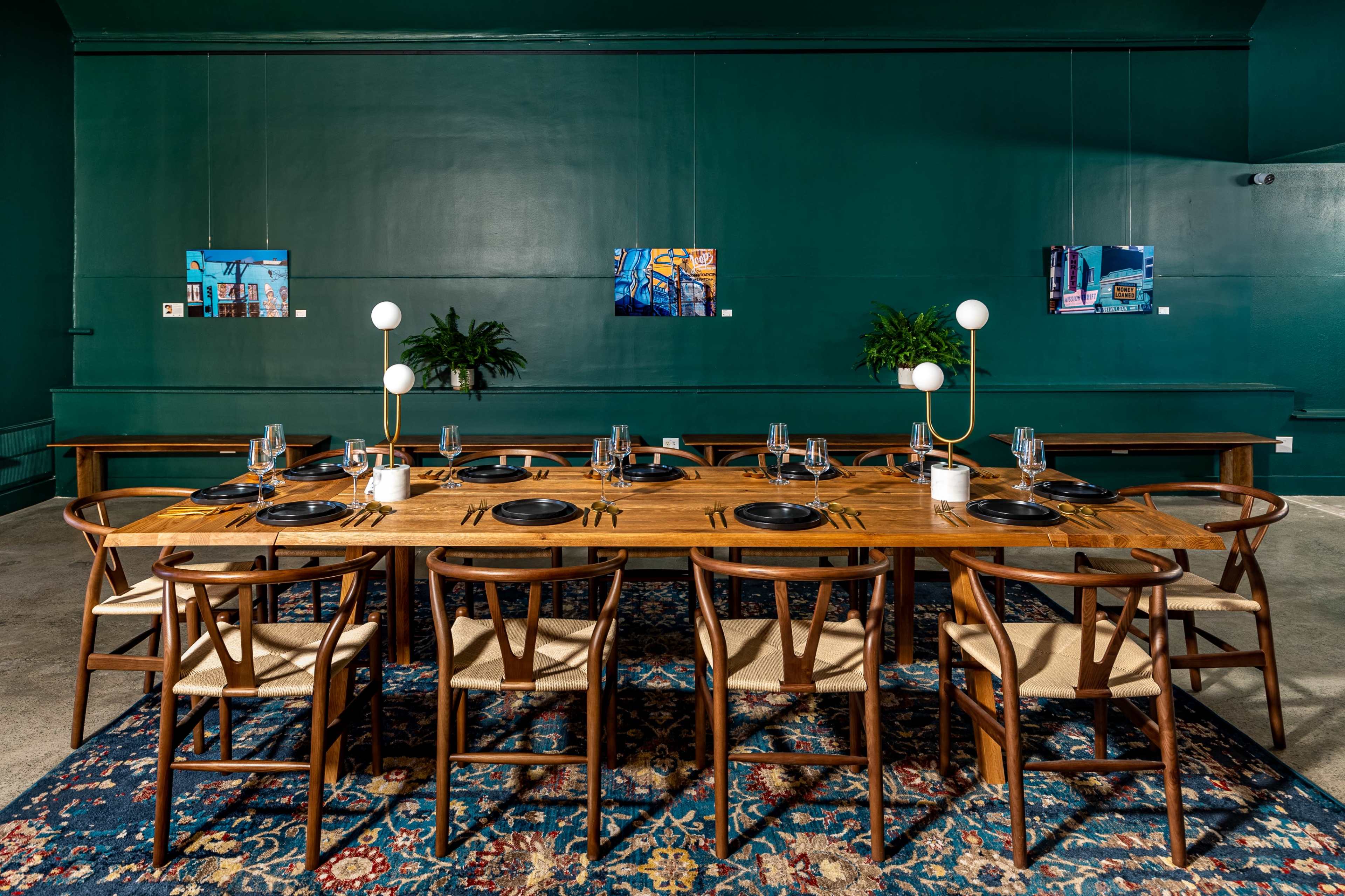 A large wooden dining table is set with plates and glasses, surrounded by chairs, in a room with dark green walls and artwork displayed.