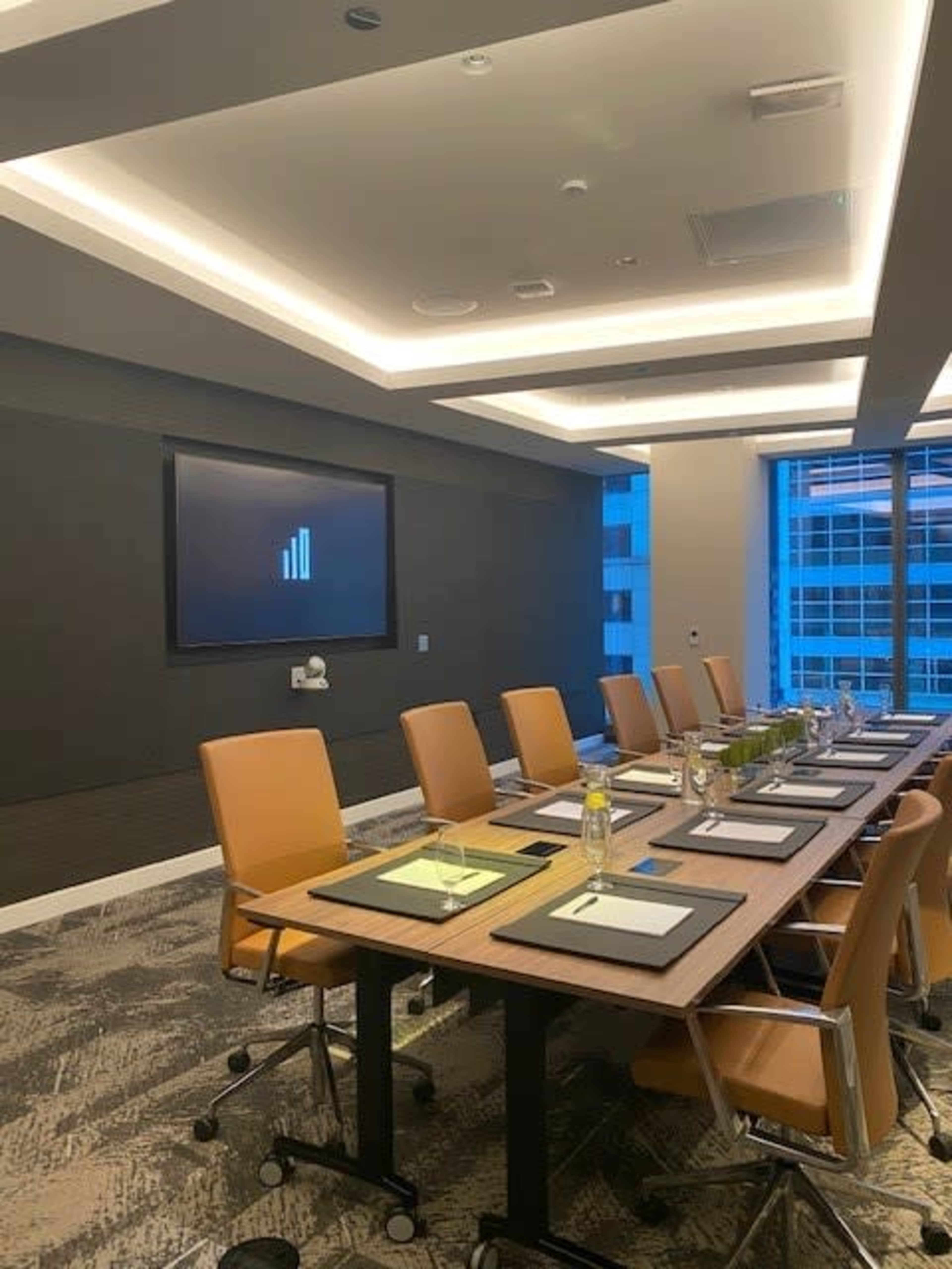 A modern conference room features a long table with ten chairs, a large screen displaying a signal icon, and windows overlooking a cityscape.
