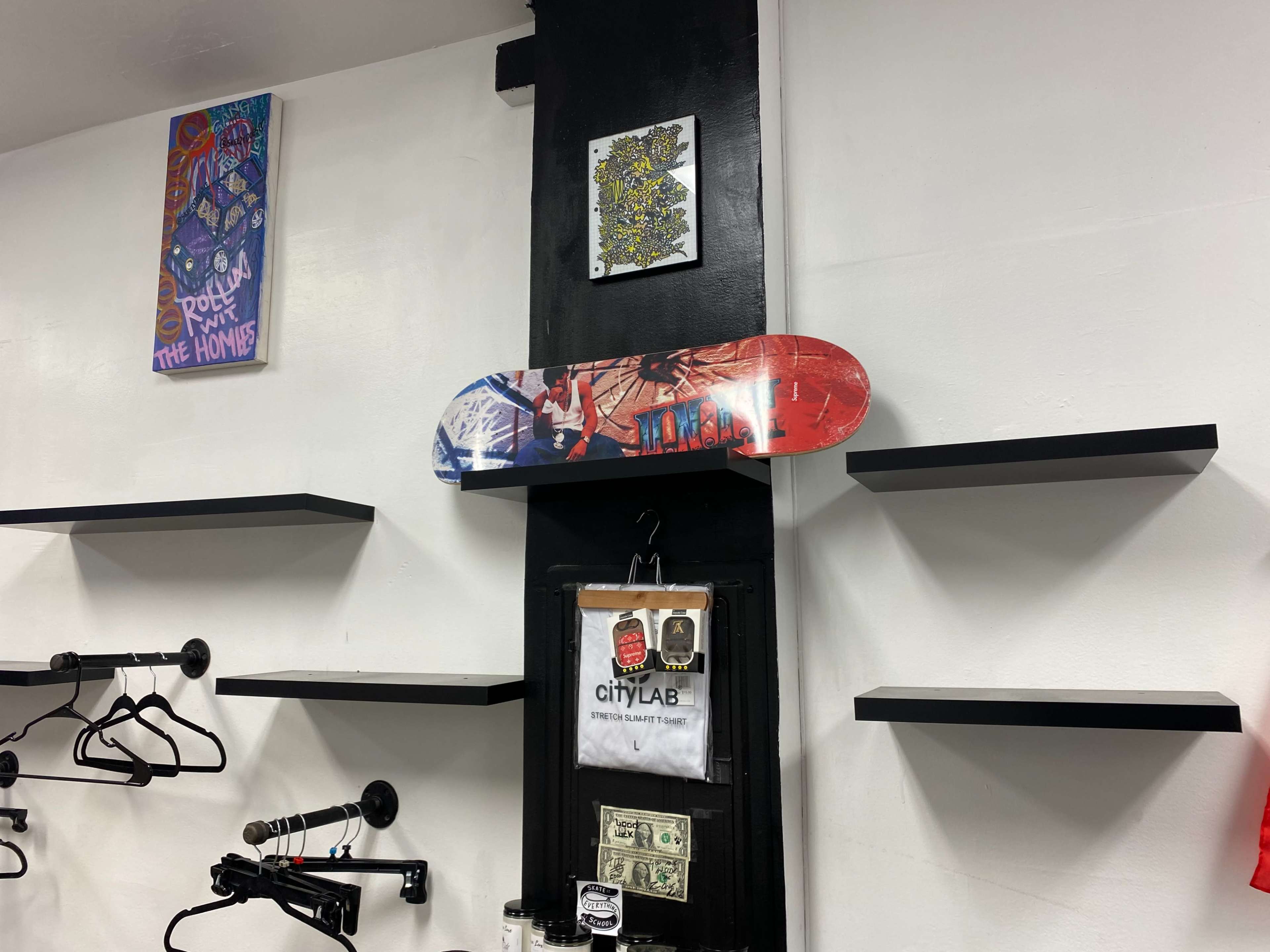 A wall display featuring a colorful skateboard mounted above two black shelves, alongside various framed artworks and items.