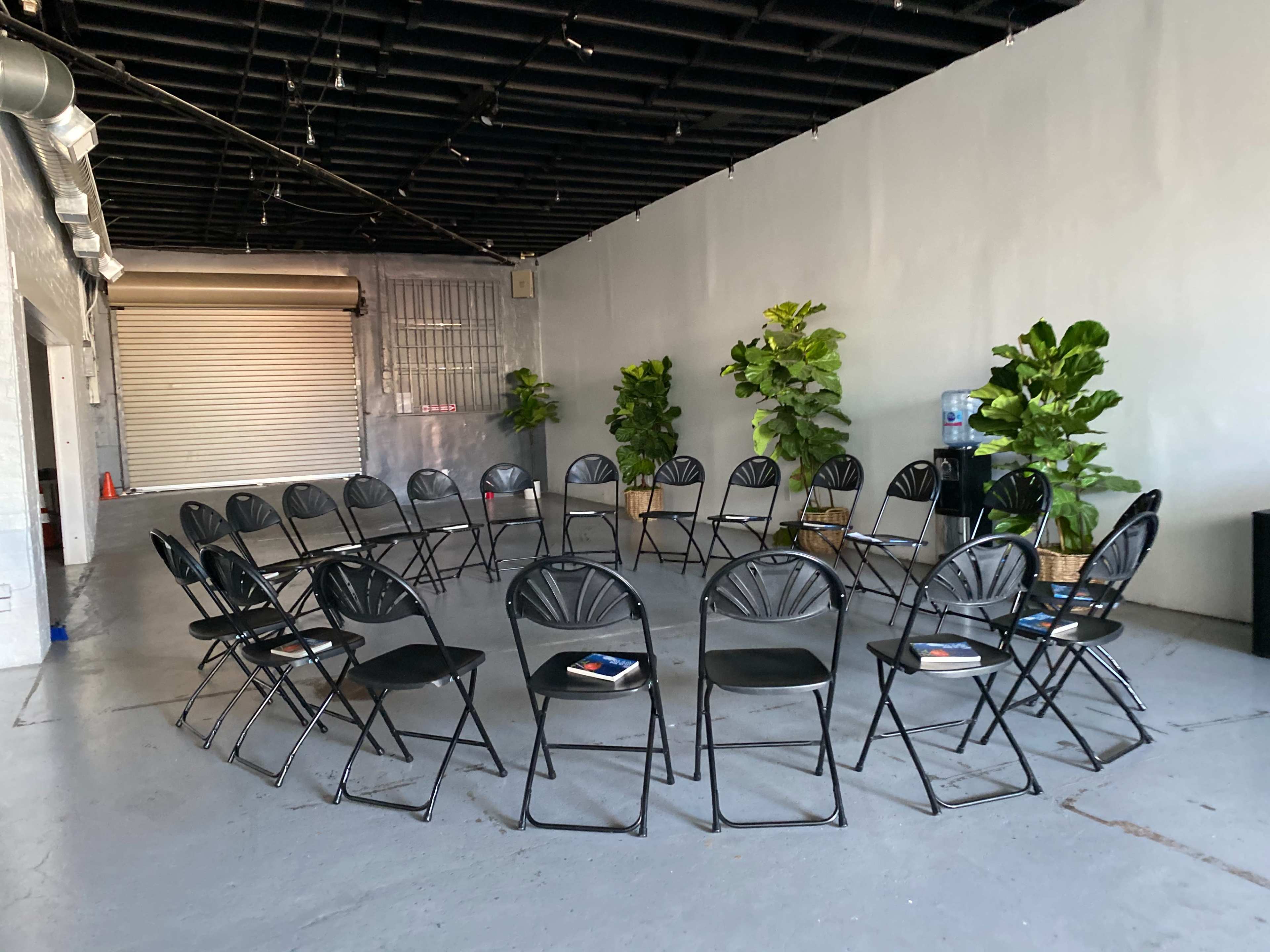 A room arranged with a circular seating layout of black folding chairs around potted plants and a water cooler.