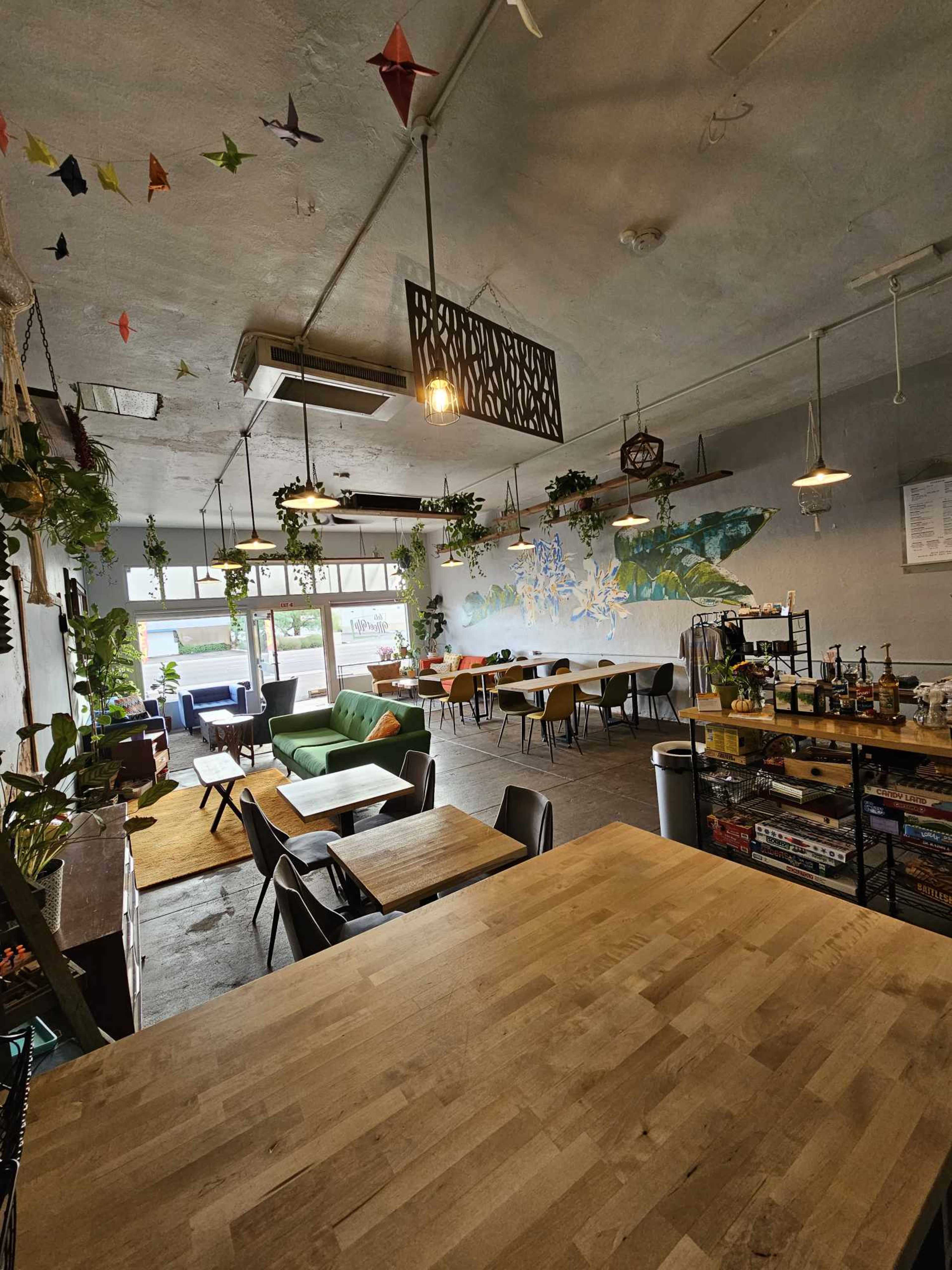 A spacious cafe with various seating areas, plants hanging from the ceiling, and a colorful mural on the wall.