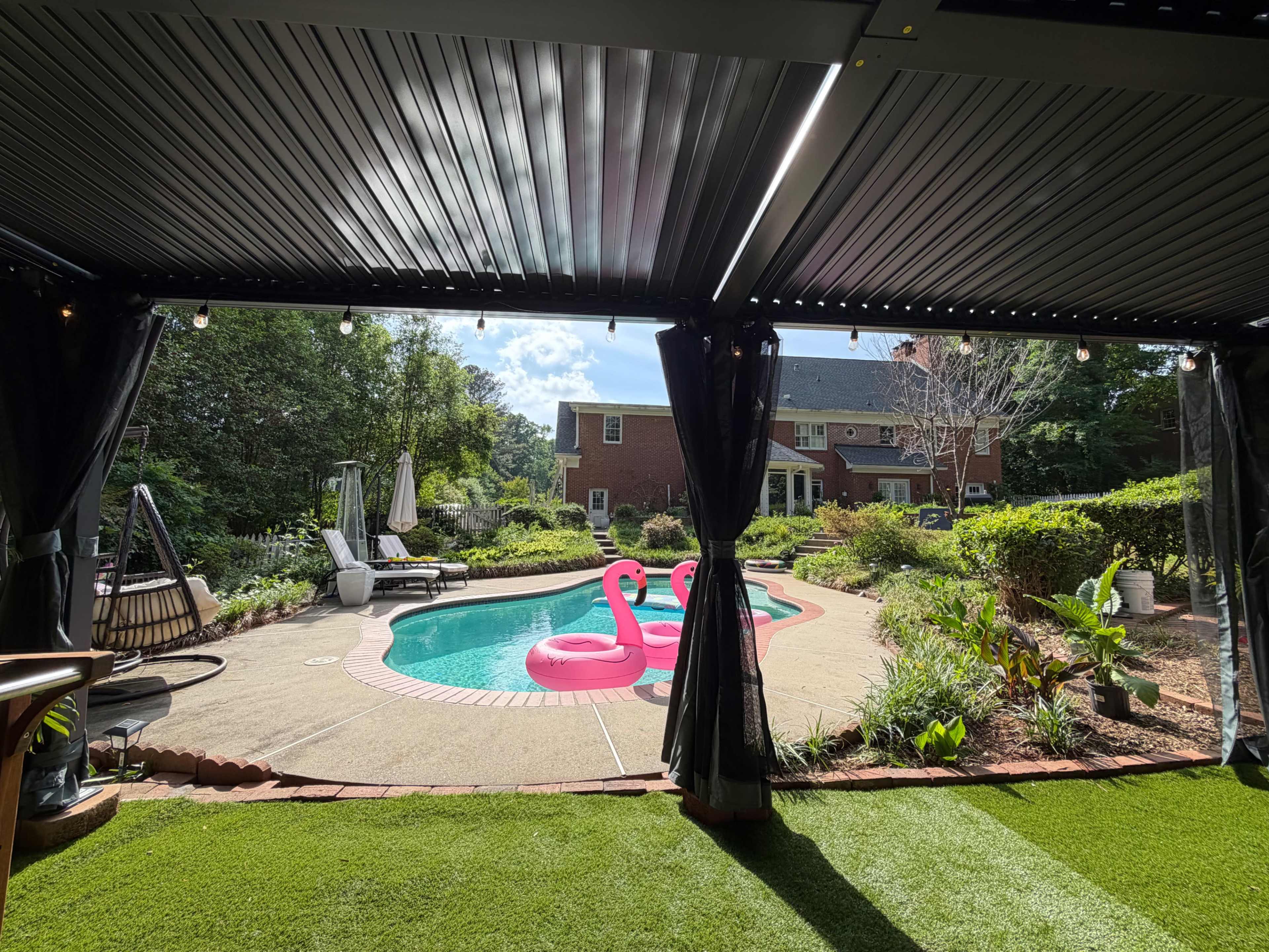 The image shows a backyard with a swimming pool surrounded by landscaped greenery, two pink inflatable flamingos, and a house in the background.