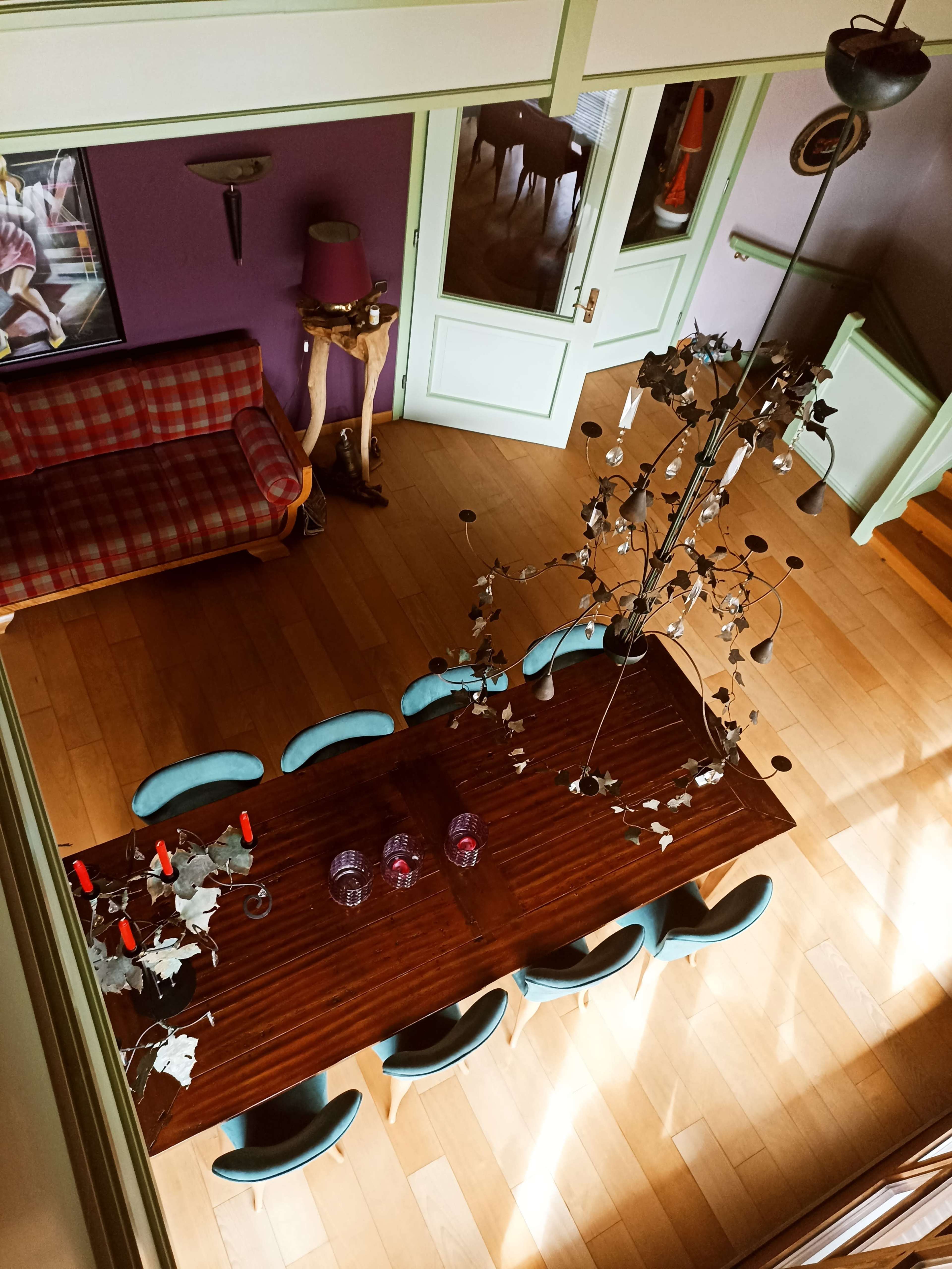 The image shows a spacious dining area with a large wooden table surrounded by eight blue chairs, a decorative chandelier, and a view of a sofa and staircase in the background.