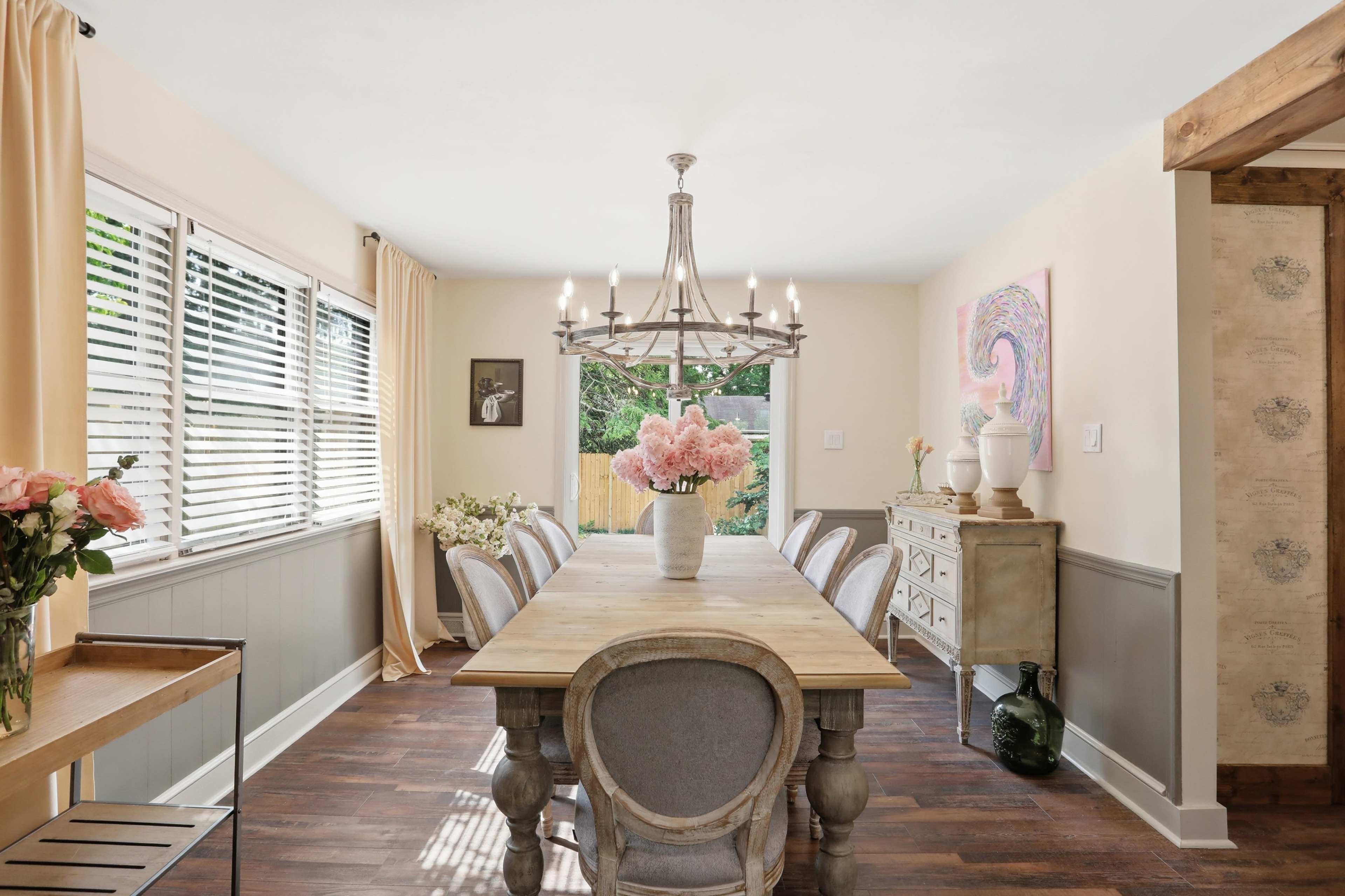 The dining room features a long wooden table surrounded by upholstered chairs, with large windows allowing natural light and adorned with floral arrangements.