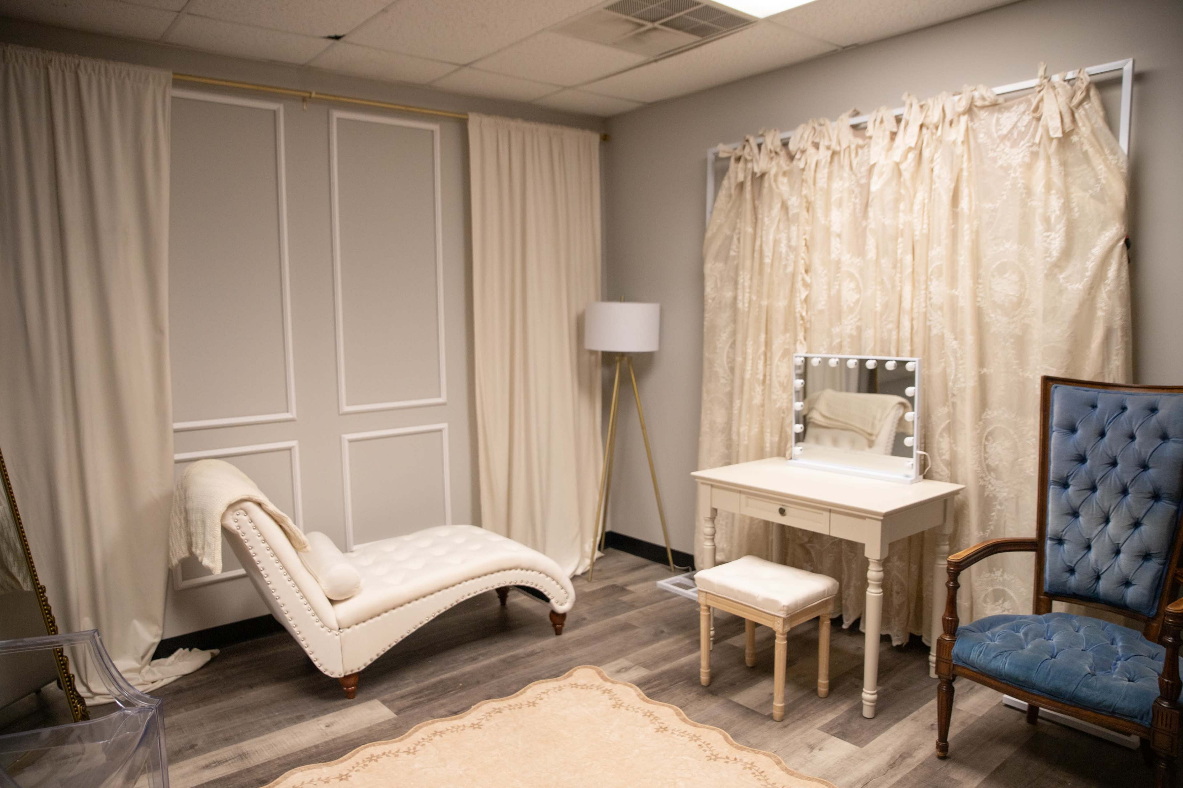 The image features a small, well-lit room with a chaise lounge, a decorative mirror on a vanity, and a blue upholstered chair, surrounded by cream-colored curtains and a patterned rug.