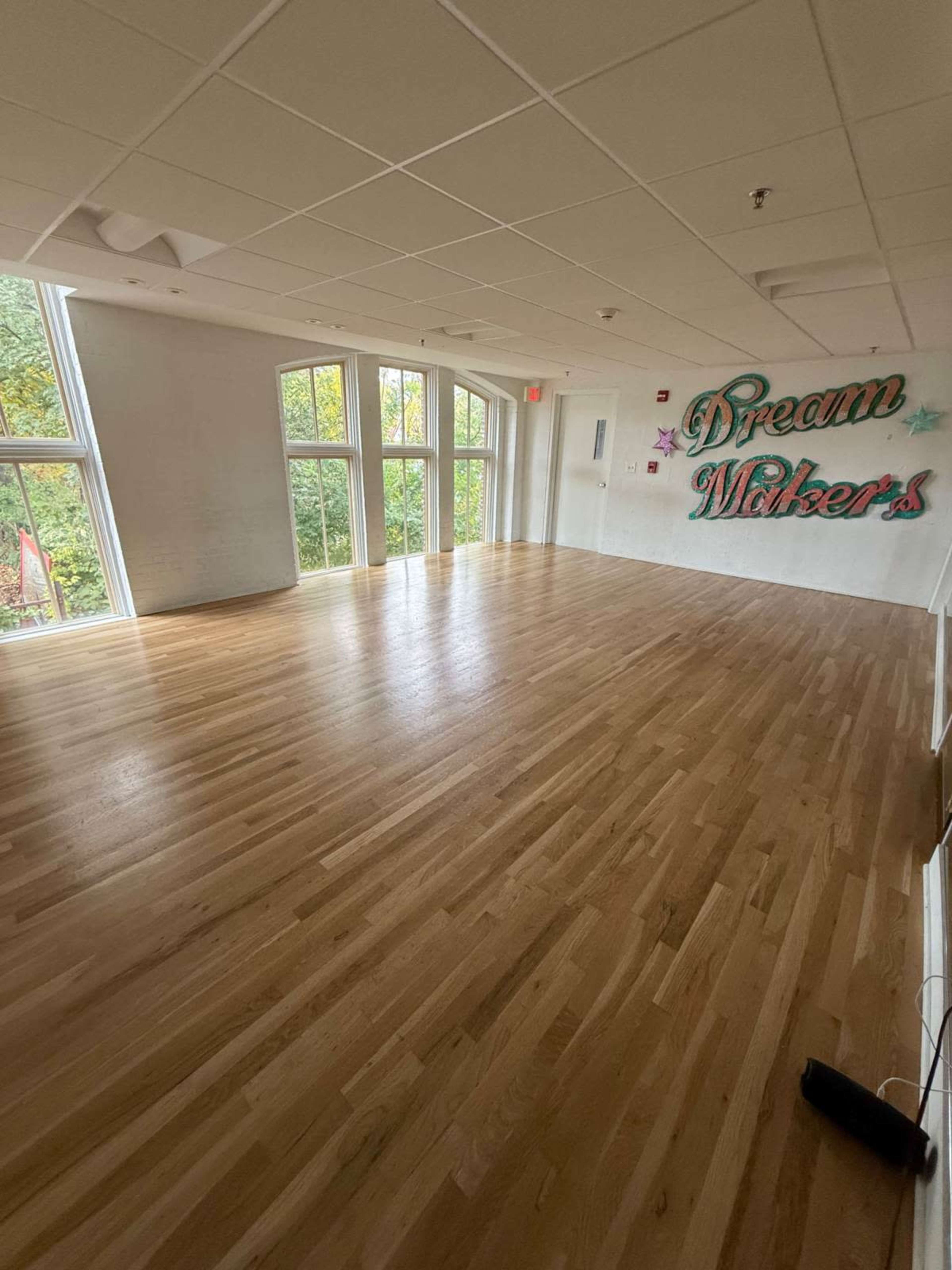 The image shows a bright, empty room with large windows and a hardwood floor, featuring the words "Dream Makers" on the wall.