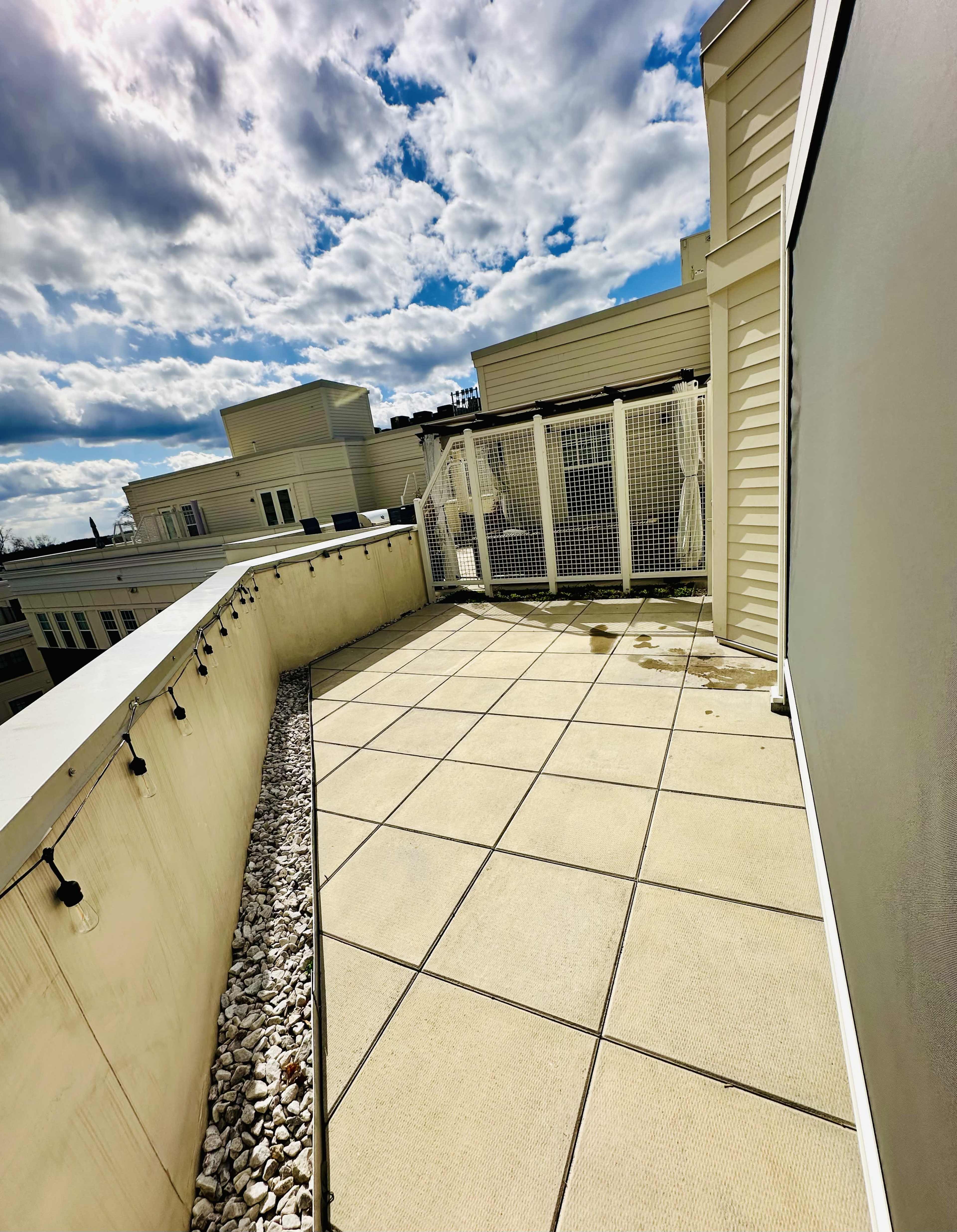 A rooftop terrace features a tiled floor, surrounded by low walls and a wire fence, under a cloudy sky.