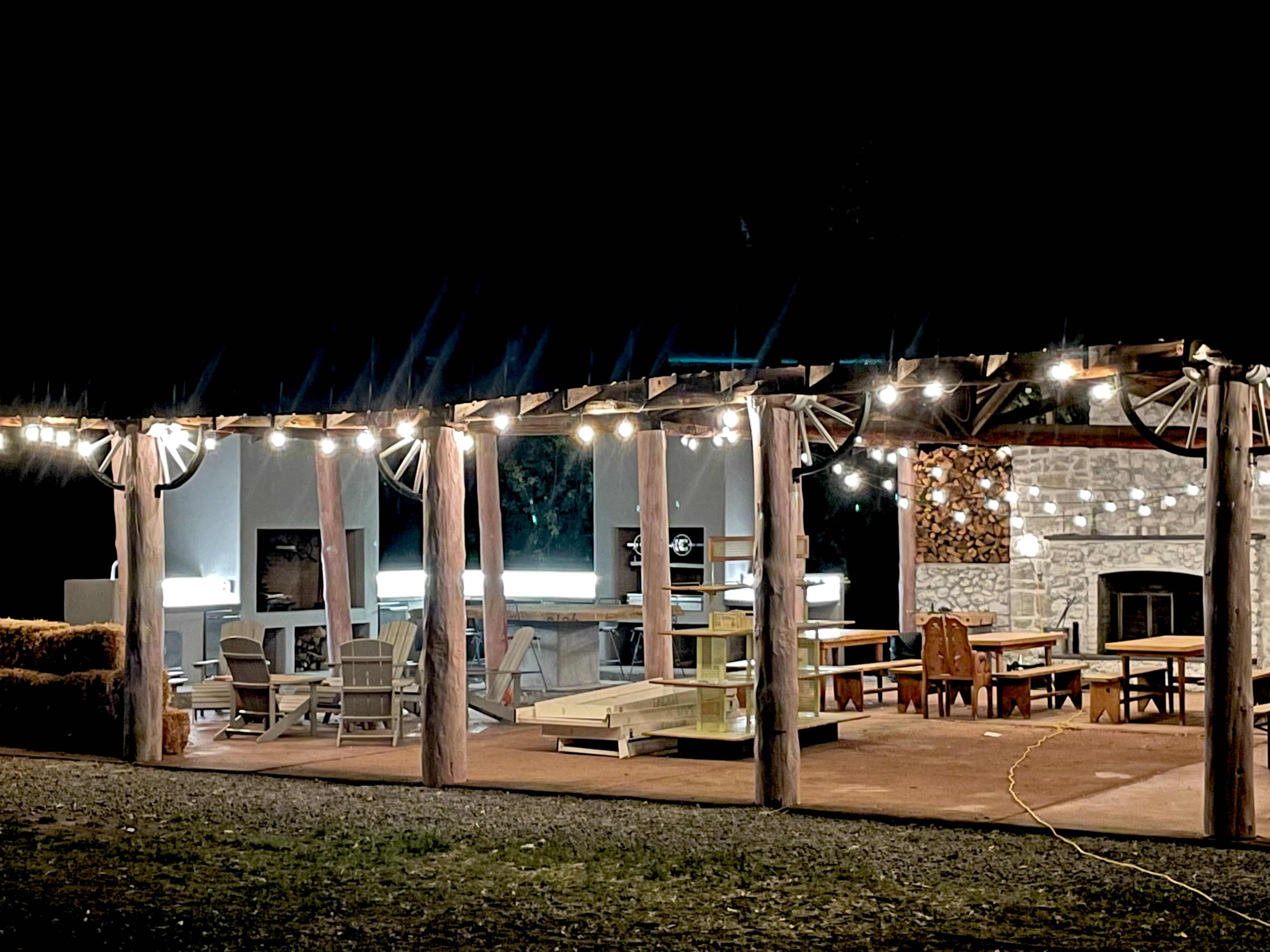 A well-lit outdoor patio features a variety of seating options and a stone fireplace under a wooden pergola at night.