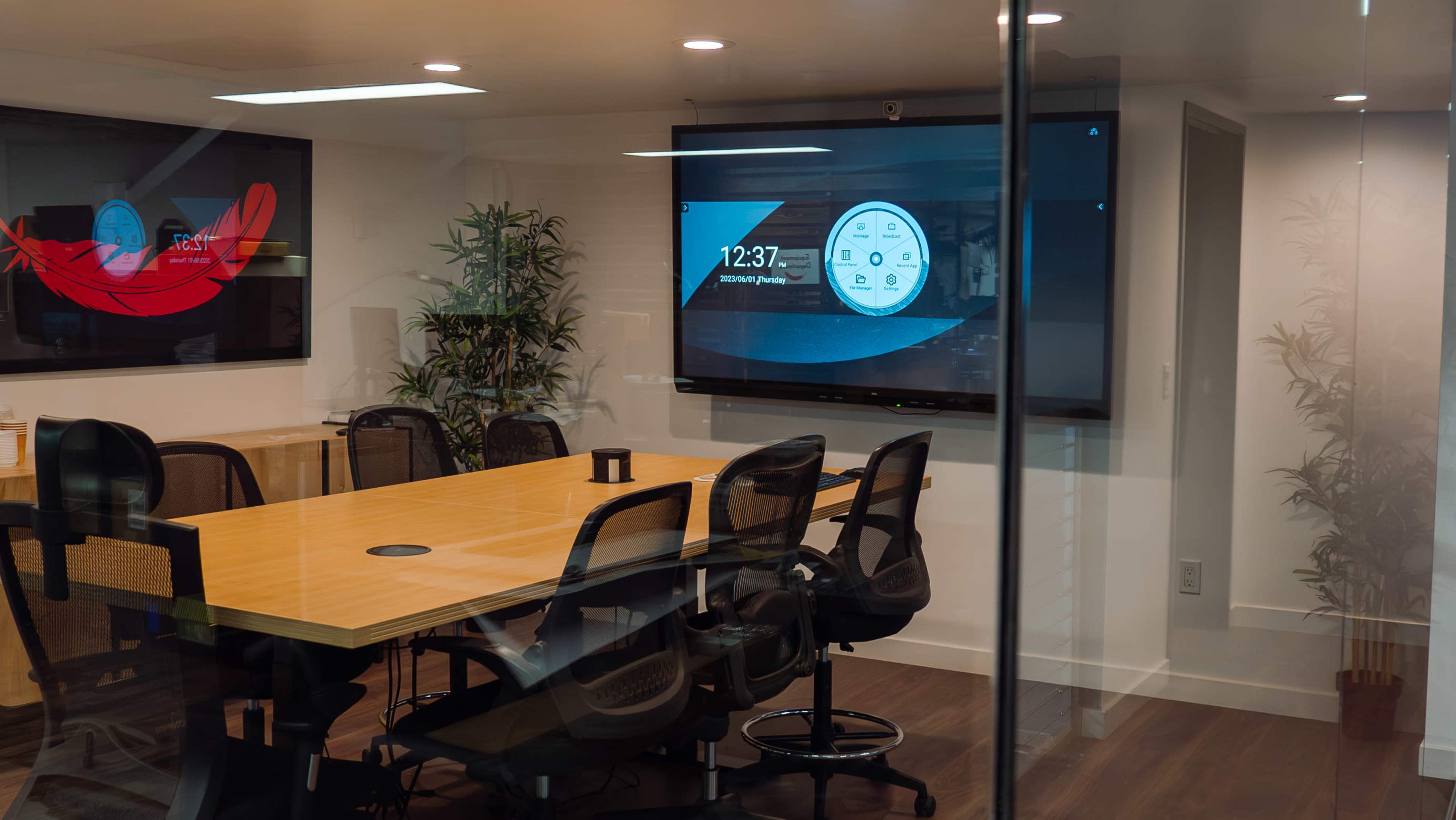 A conference room features a long wooden table surrounded by chairs, with digital screens displaying the time and a circular interface on the walls.