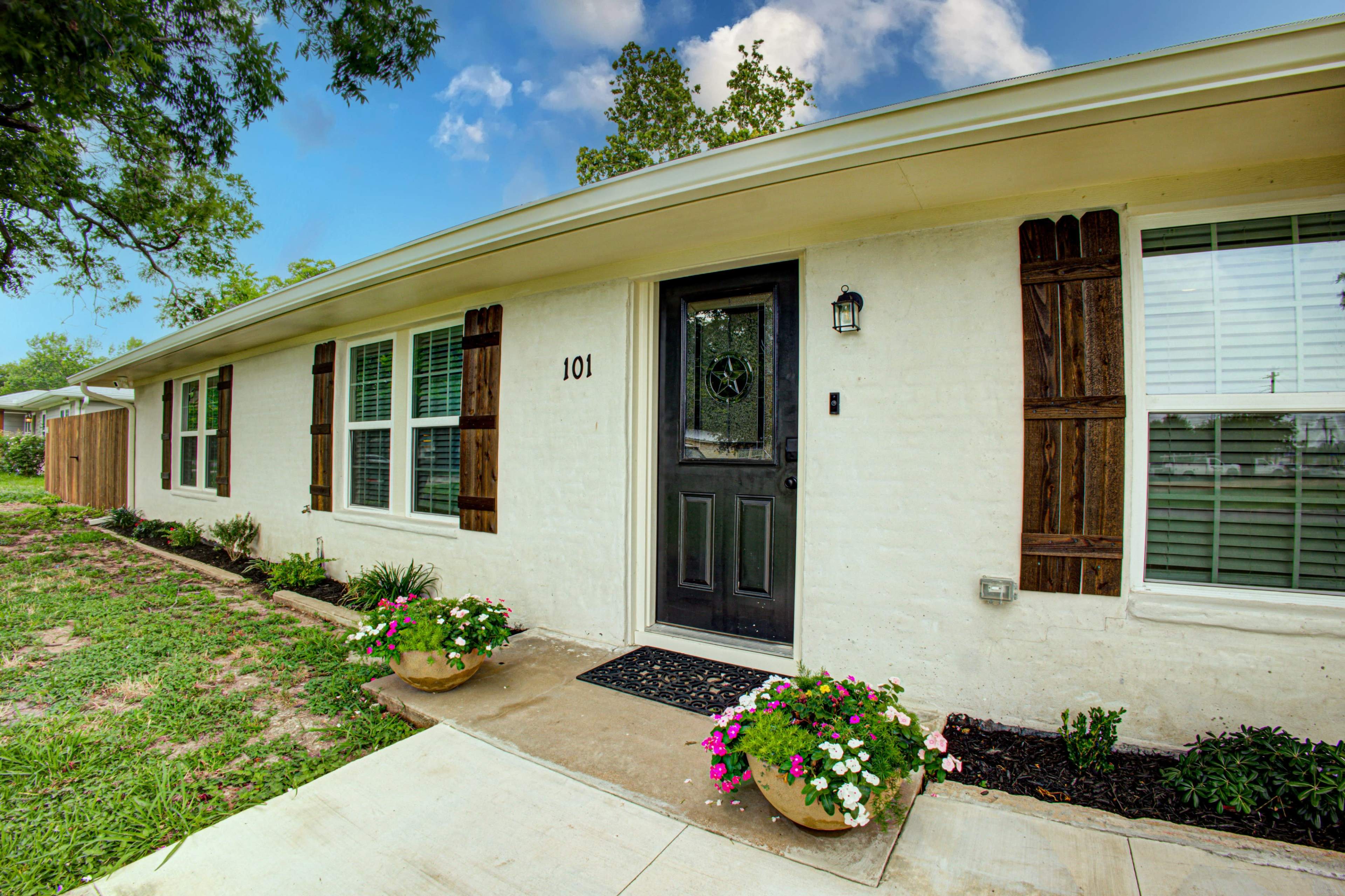 A single-story residence with a black front door, white exterior walls, wooden shutters, and flower pots filled with colorful flowers by the entrance.