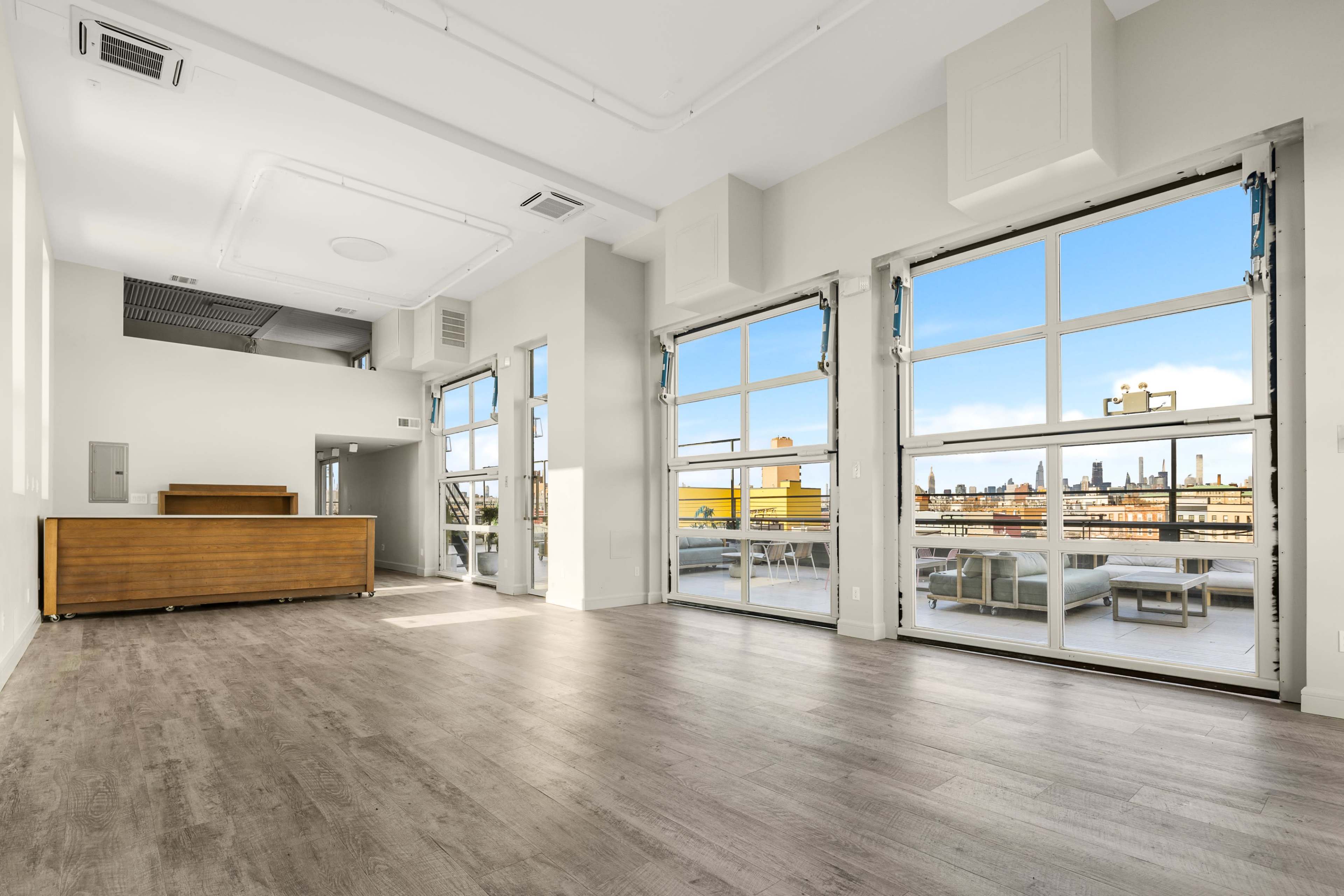 A spacious, modern interior with large windows offering a view of a city skyline and light-colored wooden flooring.