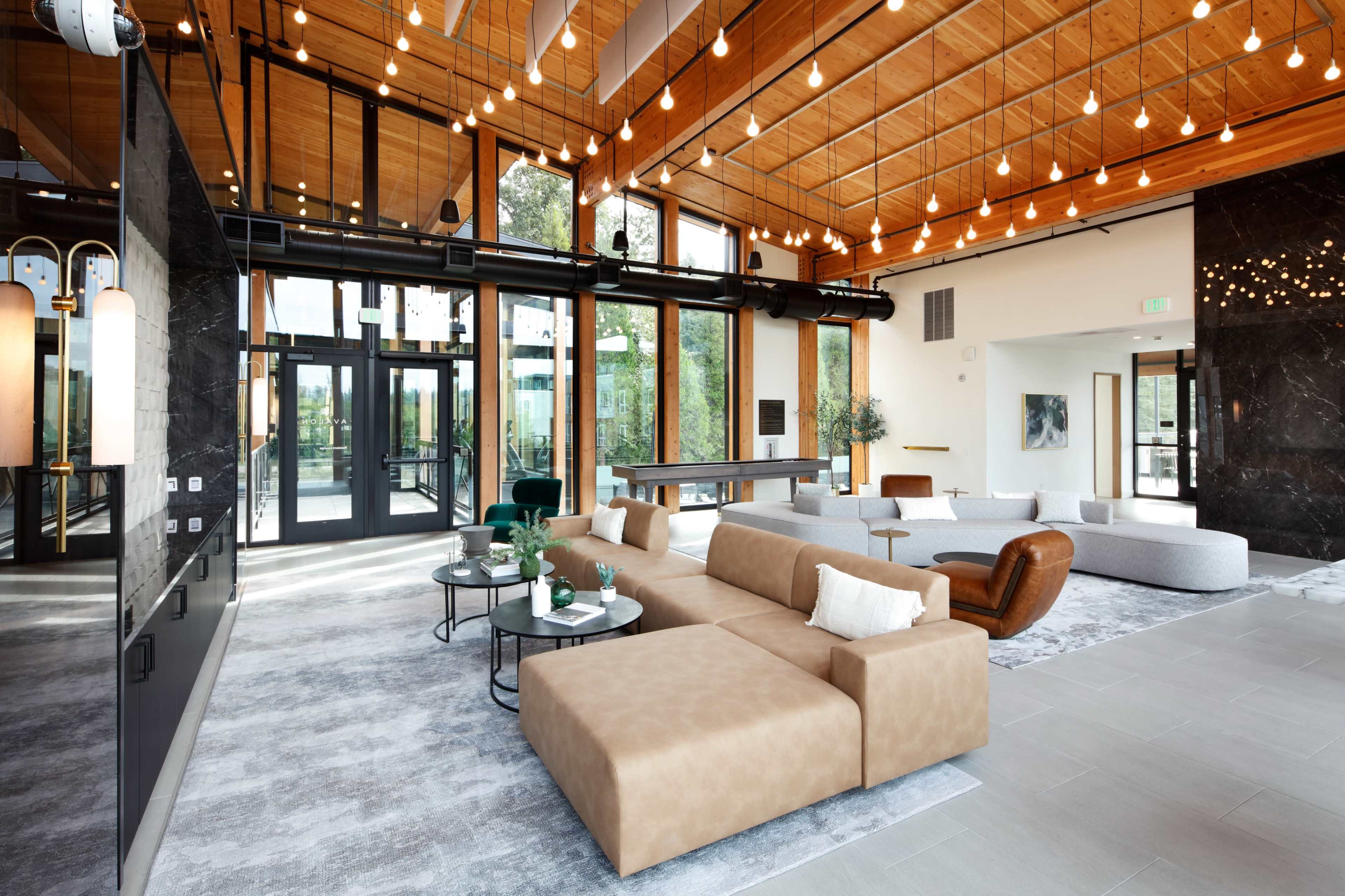 The image shows a modern lounge area featuring a combination of wooden ceilings, large windows, and contemporary furniture arrangements.