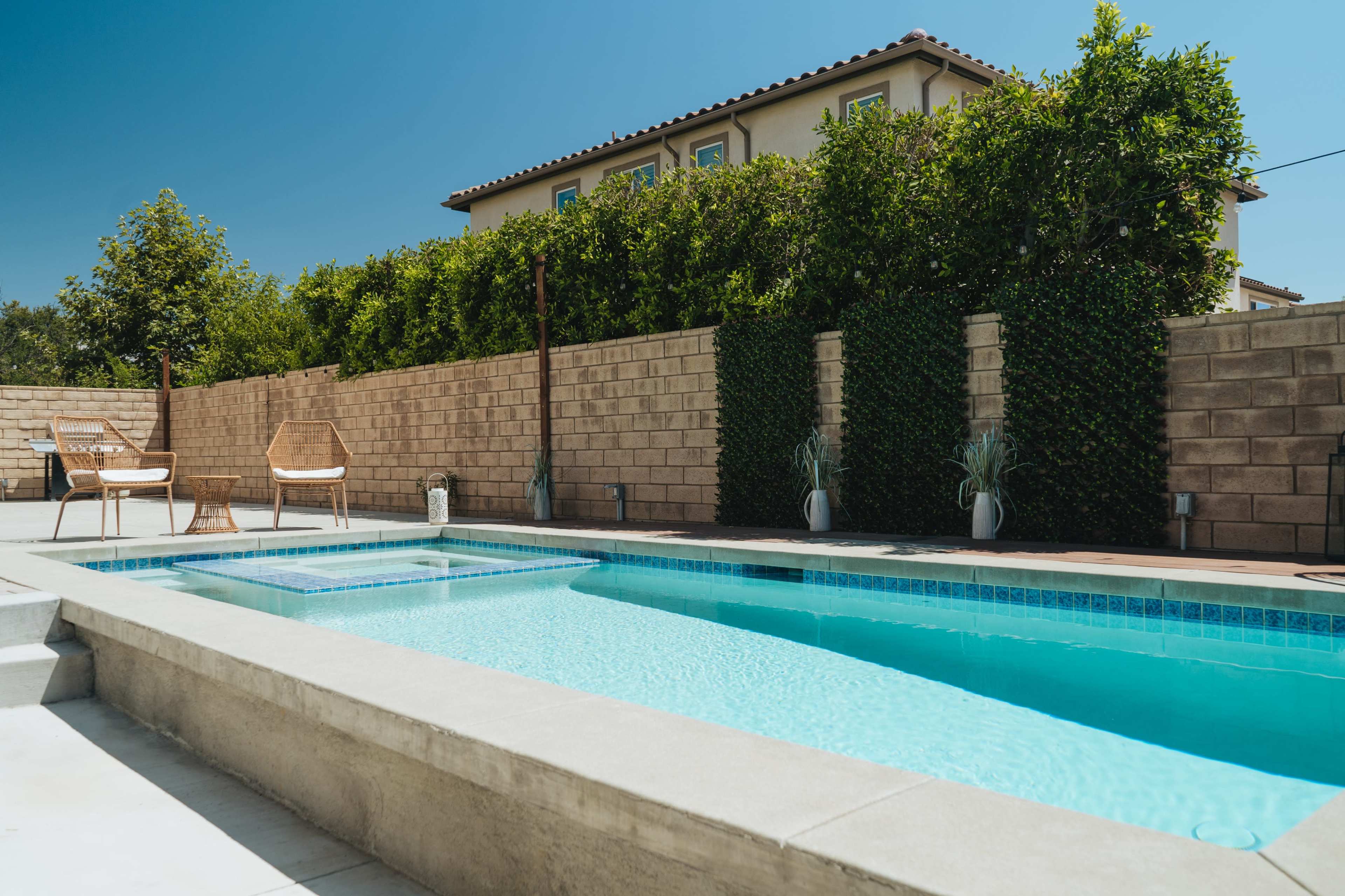 A clear swimming pool is situated beside a neatly landscaped yard with a stone wall and two chairs in the foreground.