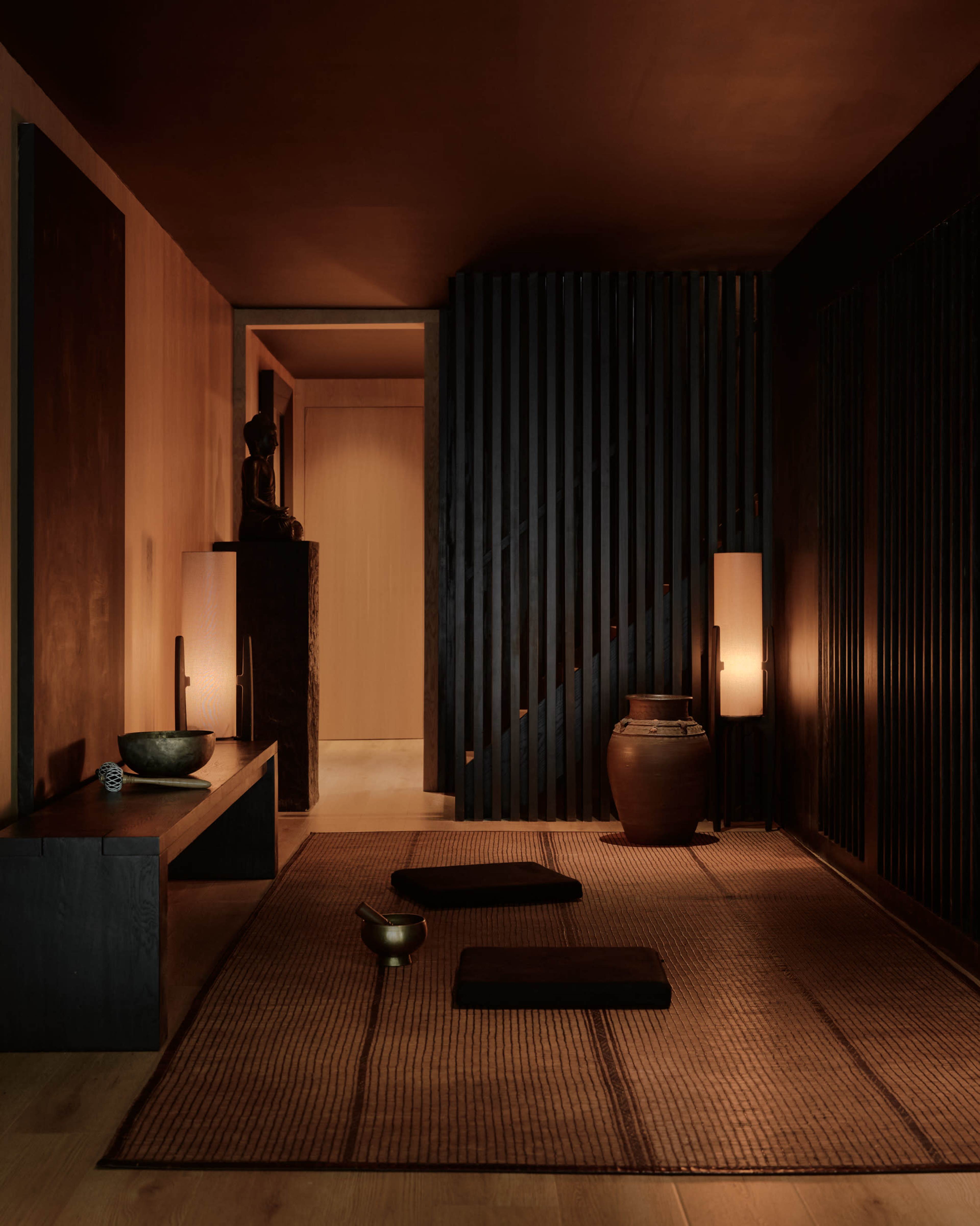 The image depicts a dimly lit interior space featuring a minimalist design with wooden elements, a statue, and softly glowing lamps.