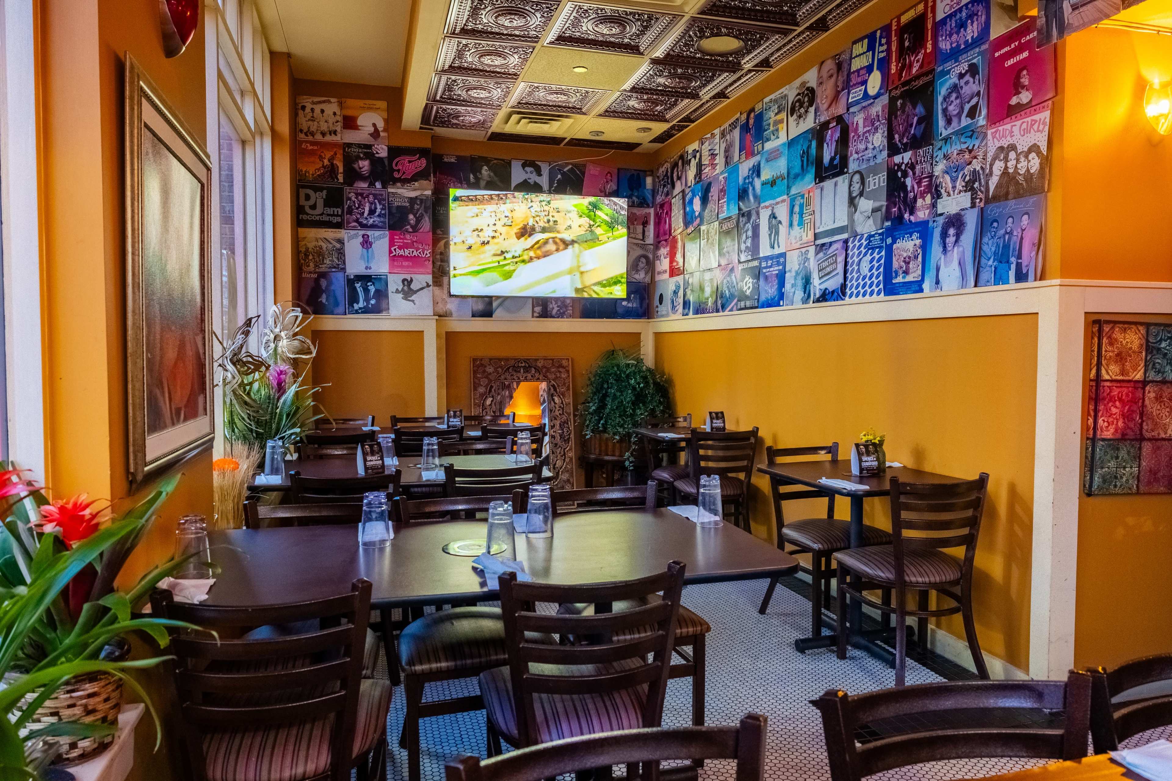 A cozy restaurant features empty tables with a wall adorned with colorful album covers and a television displaying food.