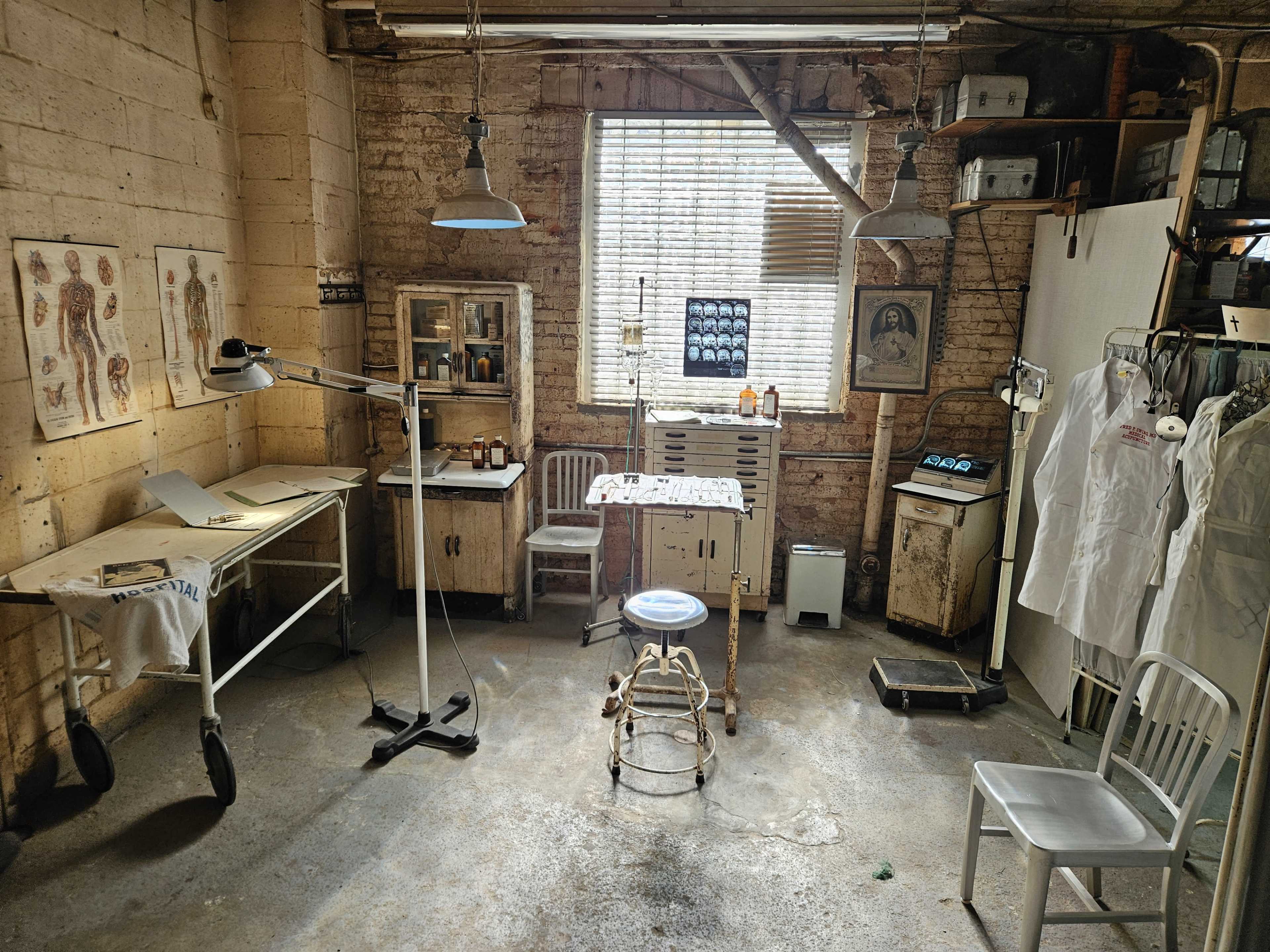 The image shows a vintage medical or examination room with various equipment, anatomical charts on the walls, and a central examination table surrounded by chairs and cabinets.