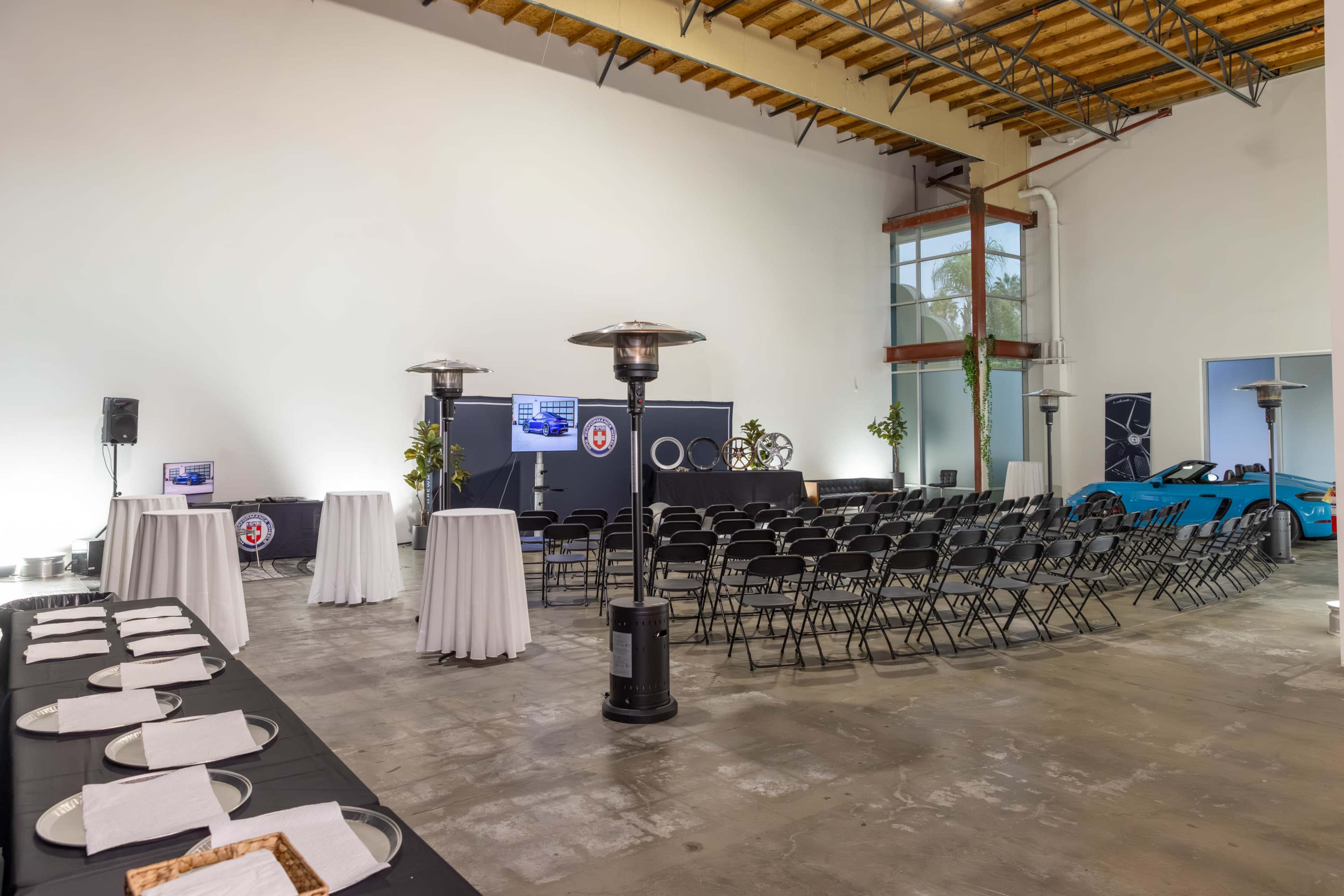 The image shows a spacious indoor event setup featuring rows of black chairs, tables with white linens, and a blue sports car parked to the side.