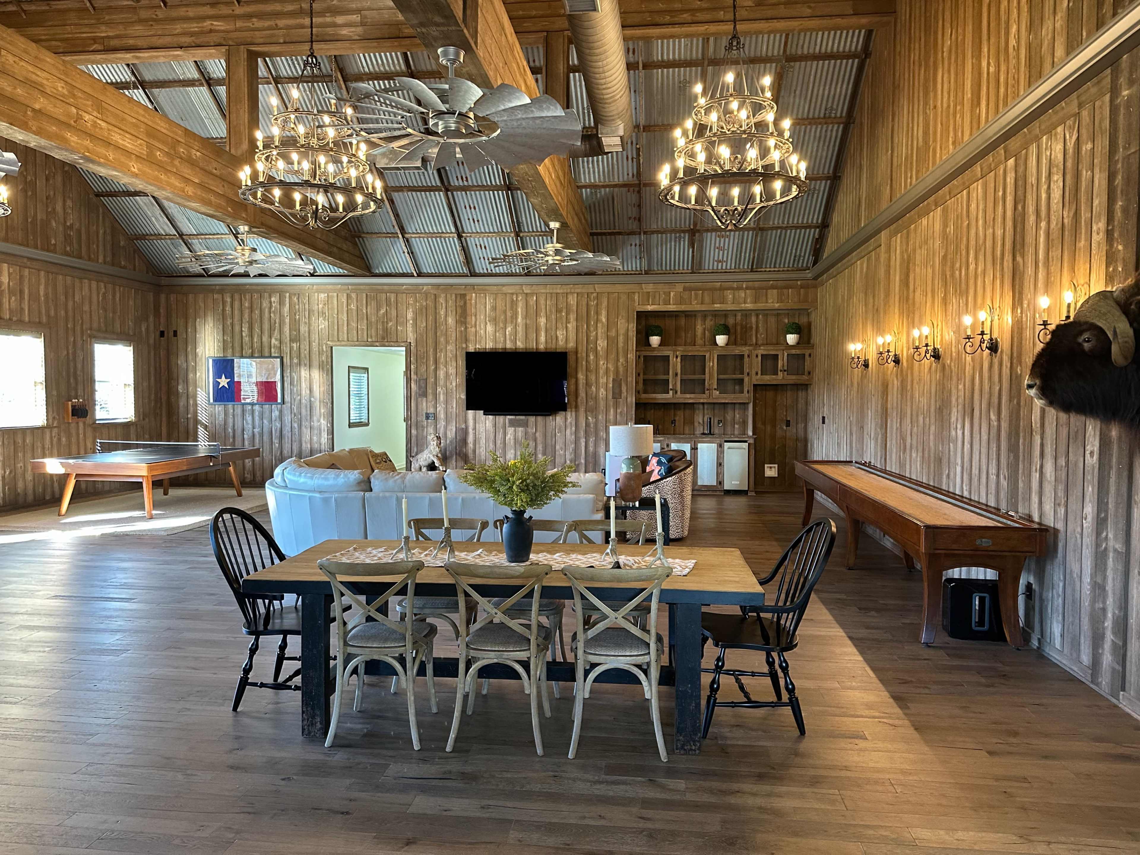 An interior of a spacious rustic event space featuring wooden walls, a long dining table with chairs, a sectional sofa, and a pool table.