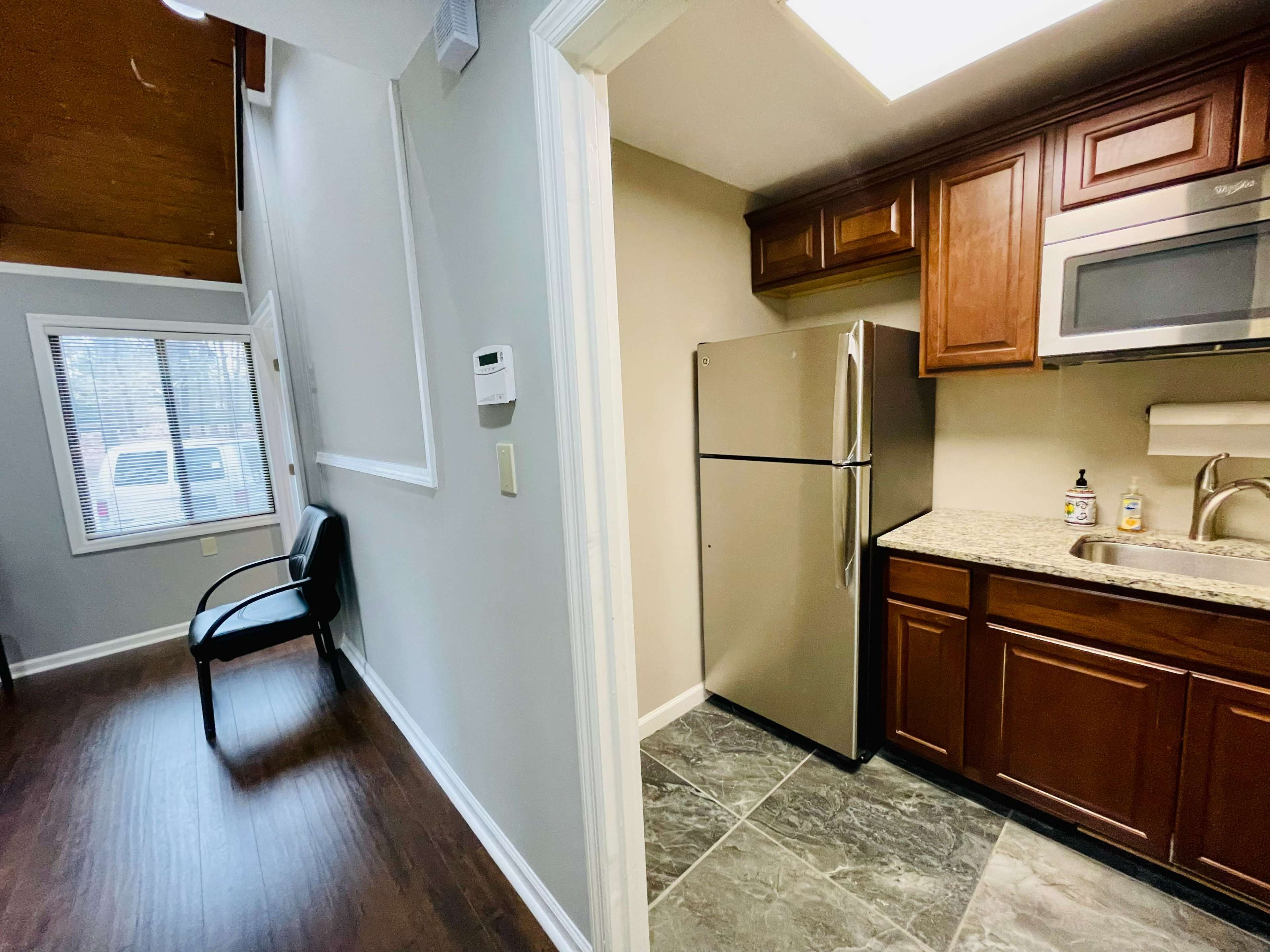 A kitchen area featuring stainless steel appliances and dark wooden cabinets, adjacent to a small hallway with a chair and a window.