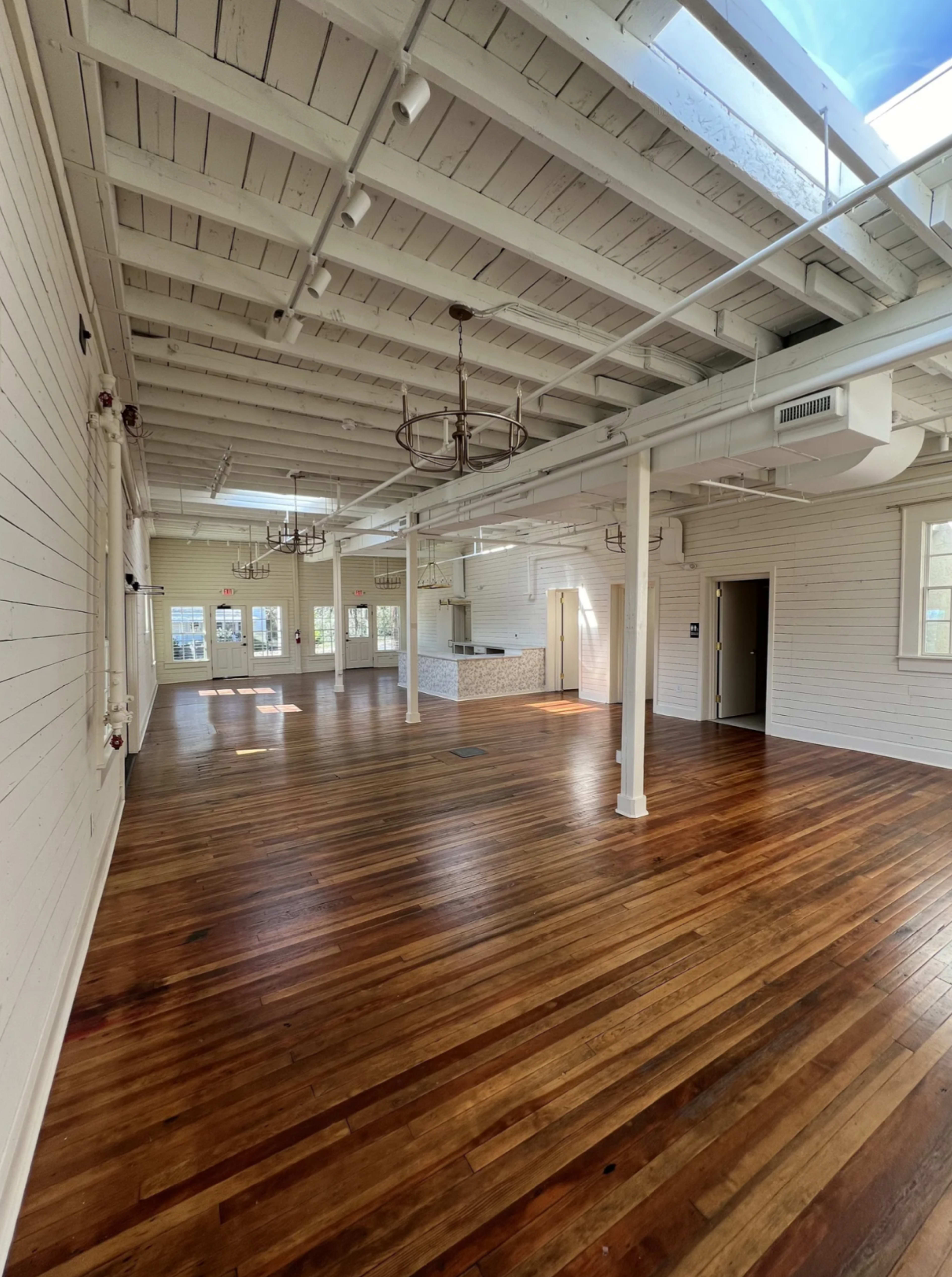 The image shows a spacious, empty room with wooden floors and white walls, featuring exposed beams and large windows.