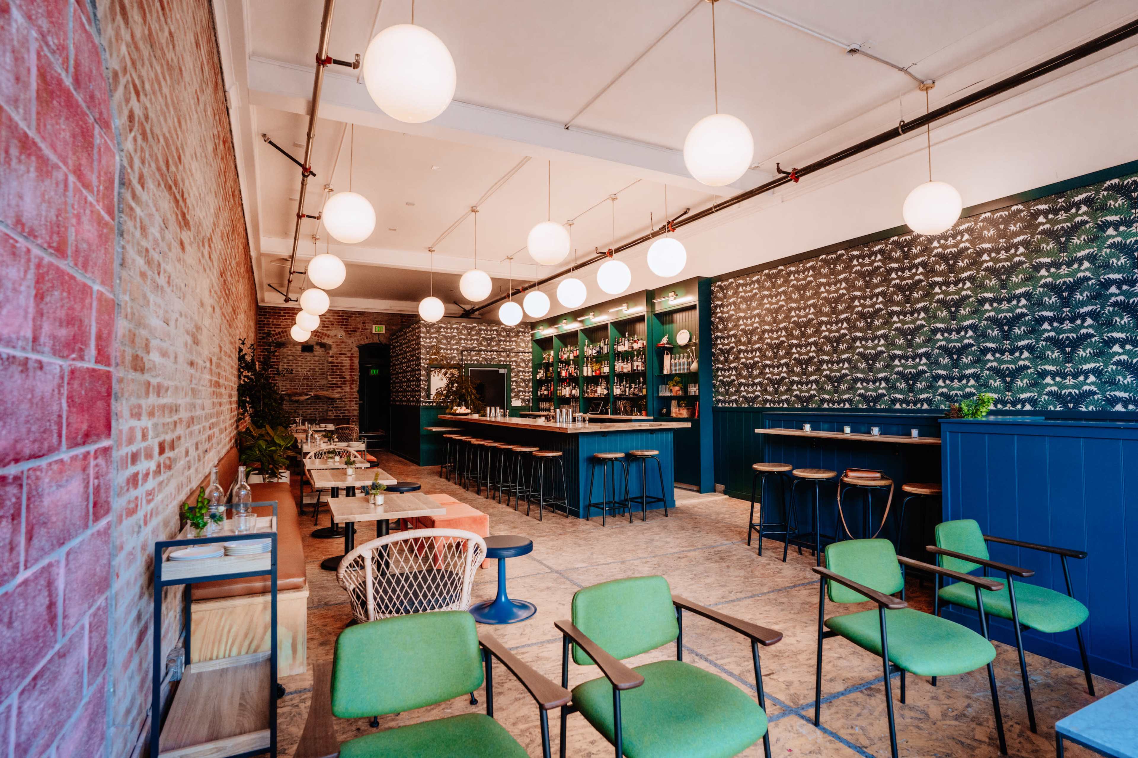 A modern bar interior features green chairs, round pendant lights, and a patterned wall with shelves stocked with bottles.