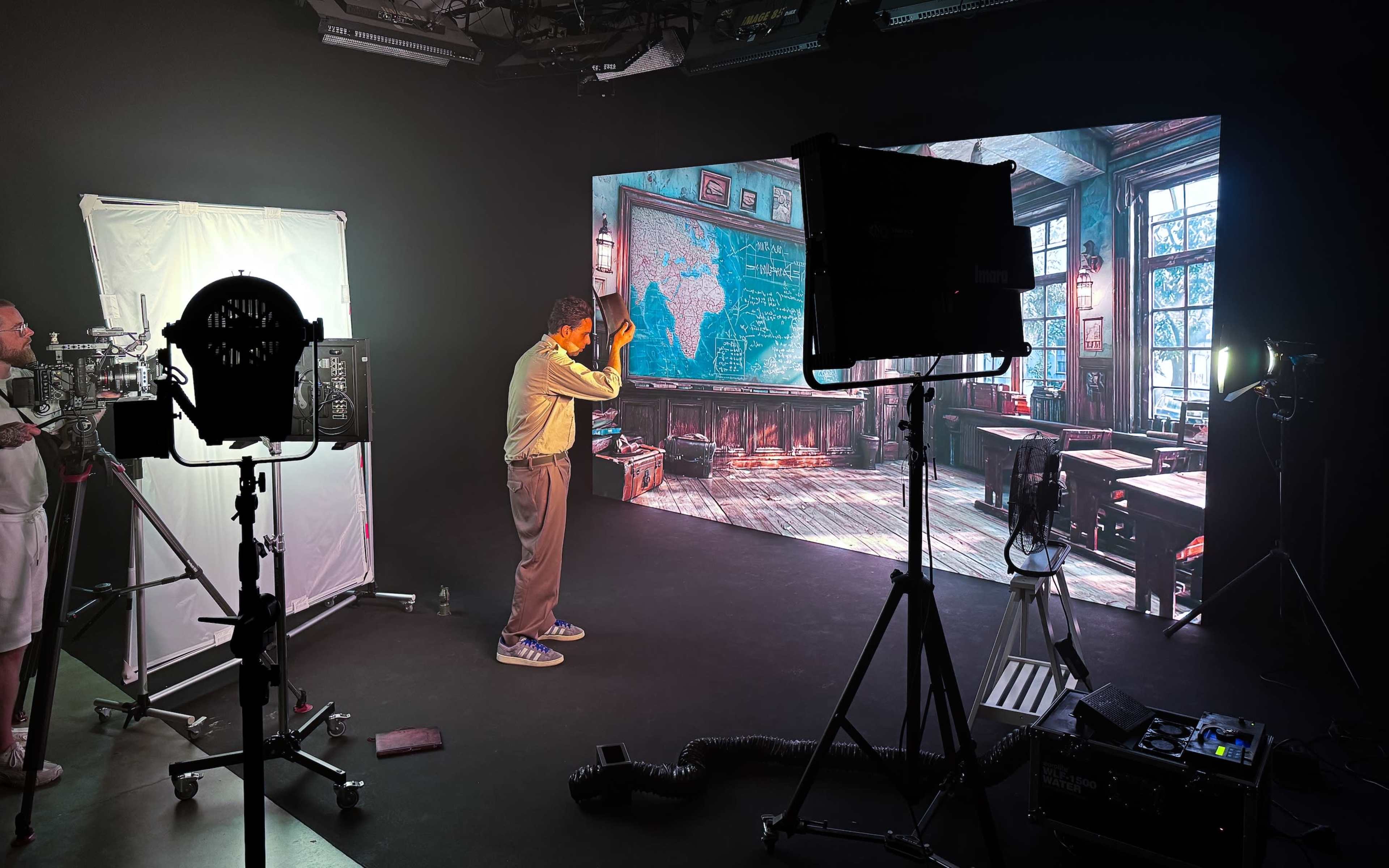 A man in a beige shirt interacts with a large digital screen displaying a classroom scene while studio lighting and equipment surround him.