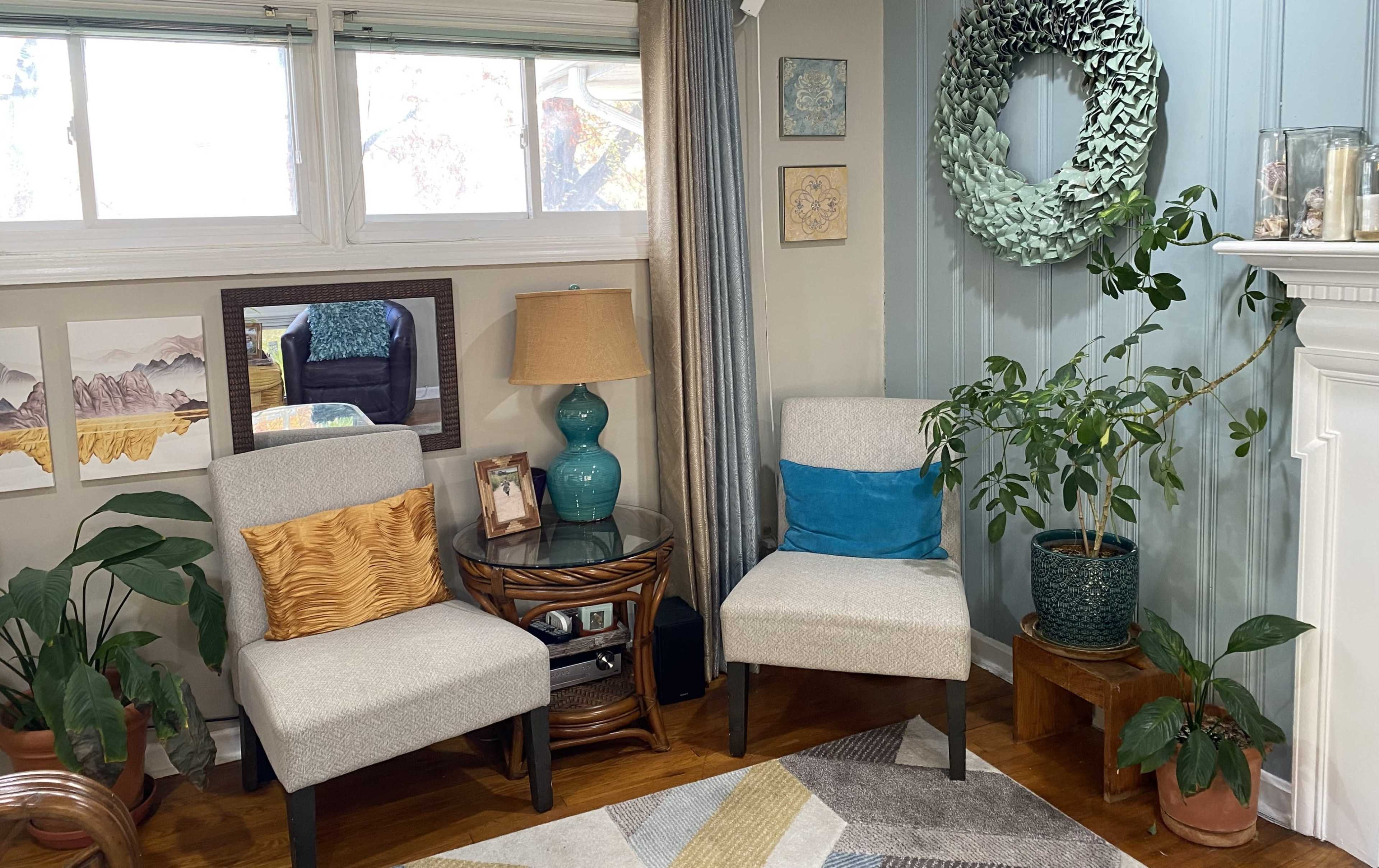 The image shows a cozy living room corner featuring two chairs, a round table with a lamp, potted plants, and wall decor.